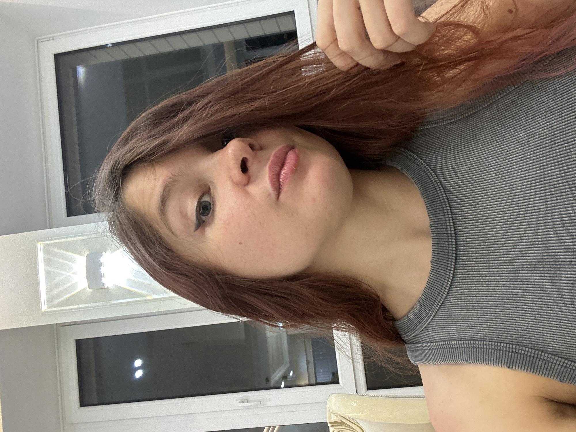 A medium, eye-level shot depicts a young woman with light skin, freckles, and long reddish-brown hair. She wears a gray striped tank top. The background features white walls and windows, one of which appears to reflect light and shadows, potentially from a city view. A white light fixture is also visible in the background. There is no information provided to suggest that this image was taken in Vladimir, Russia, or any other location.