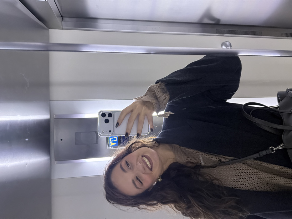 A woman takes a selfie in an elevator. She is smiling and wearing a black coat over a tan, textured sweater. Her dark, curly hair falls over her shoulder. She is holding a phone with a clear case, and her nails are painted black. A black leather bag with straps is visible. The elevator's interior features metal walls, a horizontal bar, and a panel with buttons. The image is rotated ninety degrees.