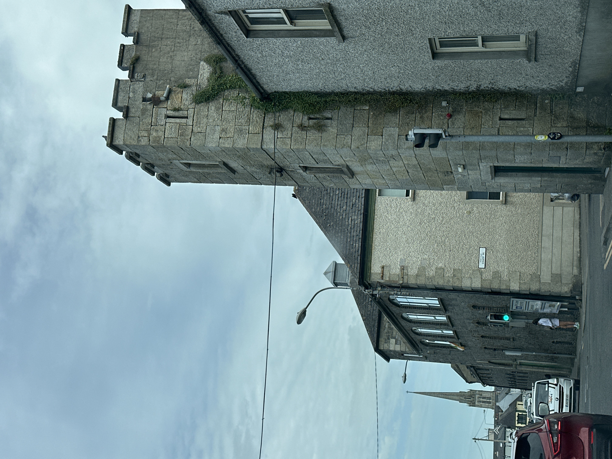 The image captures a street scene in Carlow, Ireland, with a focus on a stone tower that appears to be part of an old building, possibly a castle or historical structure. The tower is crenellated at the top and has ivy growing up its side. To the right of the tower, a gray, textured building with a window is visible. In front of the tower, a traffic light is mounted on a pole, showing a green light. A person in a white top and shorts stands near the traffic light. Behind the tower, another building with multiple arched windows is seen, and further in the distance, a church spire pierces the overcast sky. Cars are present on the street, including a red car in the foreground to the left and a white van behind it. A street sign reading "CHAFF ST" is attached to the gray building on the right. The overall atmosphere is subdued due to the cloudy sky.