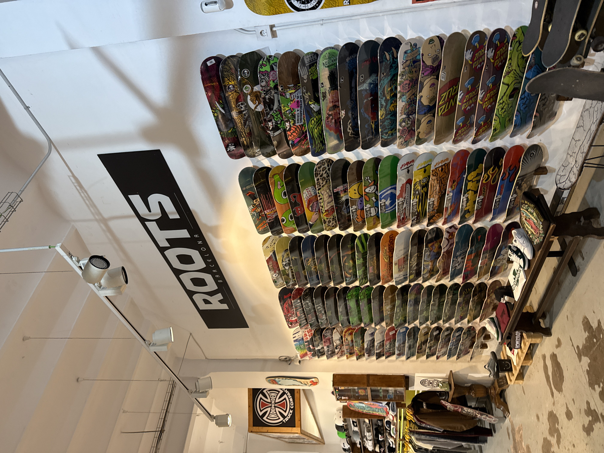 The image captures the interior of a shop called "ROOTS Barcelona" in Barcelona, Spain. The shop appears to be a skateboarding store, evidenced by the numerous skateboards mounted on the wall. The skateboards vary in design and feature a range of graphics. The store also sells related merchandise such as shoes and apparel, which are displayed on shelves and racks below the skateboards. An "INDEPENDENT TRUCK COMPANY" logo is visible. The environment is well-lit, and the walls are painted white. The floor has a spotted surface.