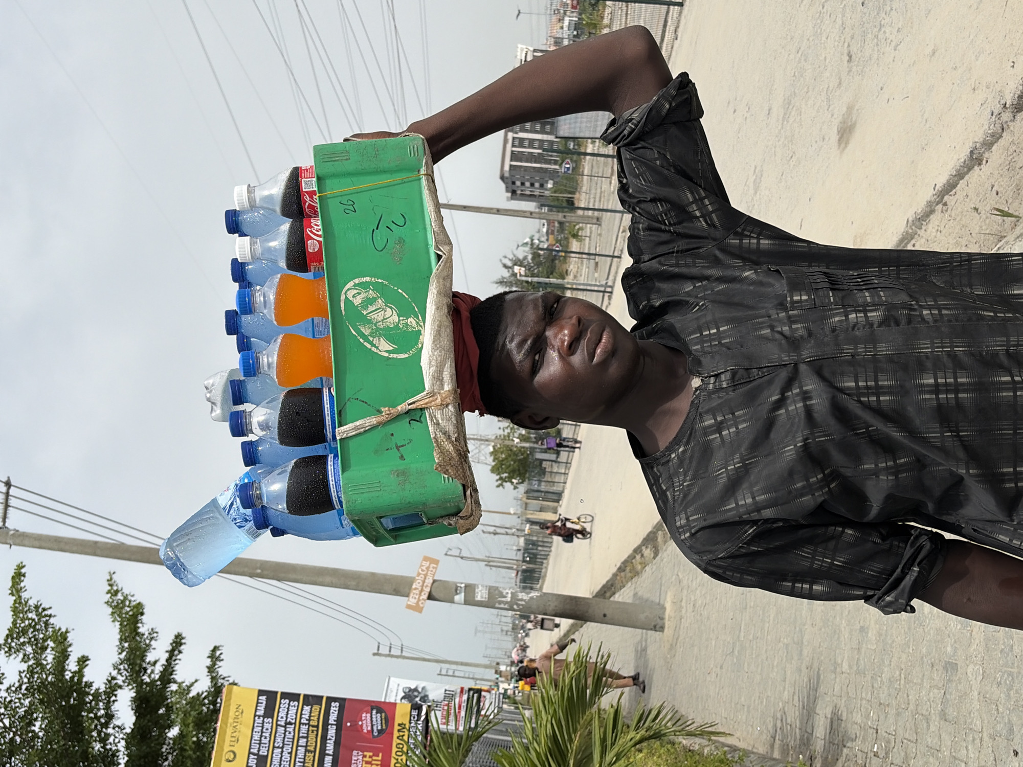 A vendor balances a green crate of bottled drinks on their head, including Coca-Cola and orange soda, on a sunlit street in Igboefon, Nigeria. The vendor is wearing a dark patterned shirt. A sign for "ELEVATION" events is visible in the background, along with power lines and buildings. Pedestrians and a cyclist are seen in the distance.