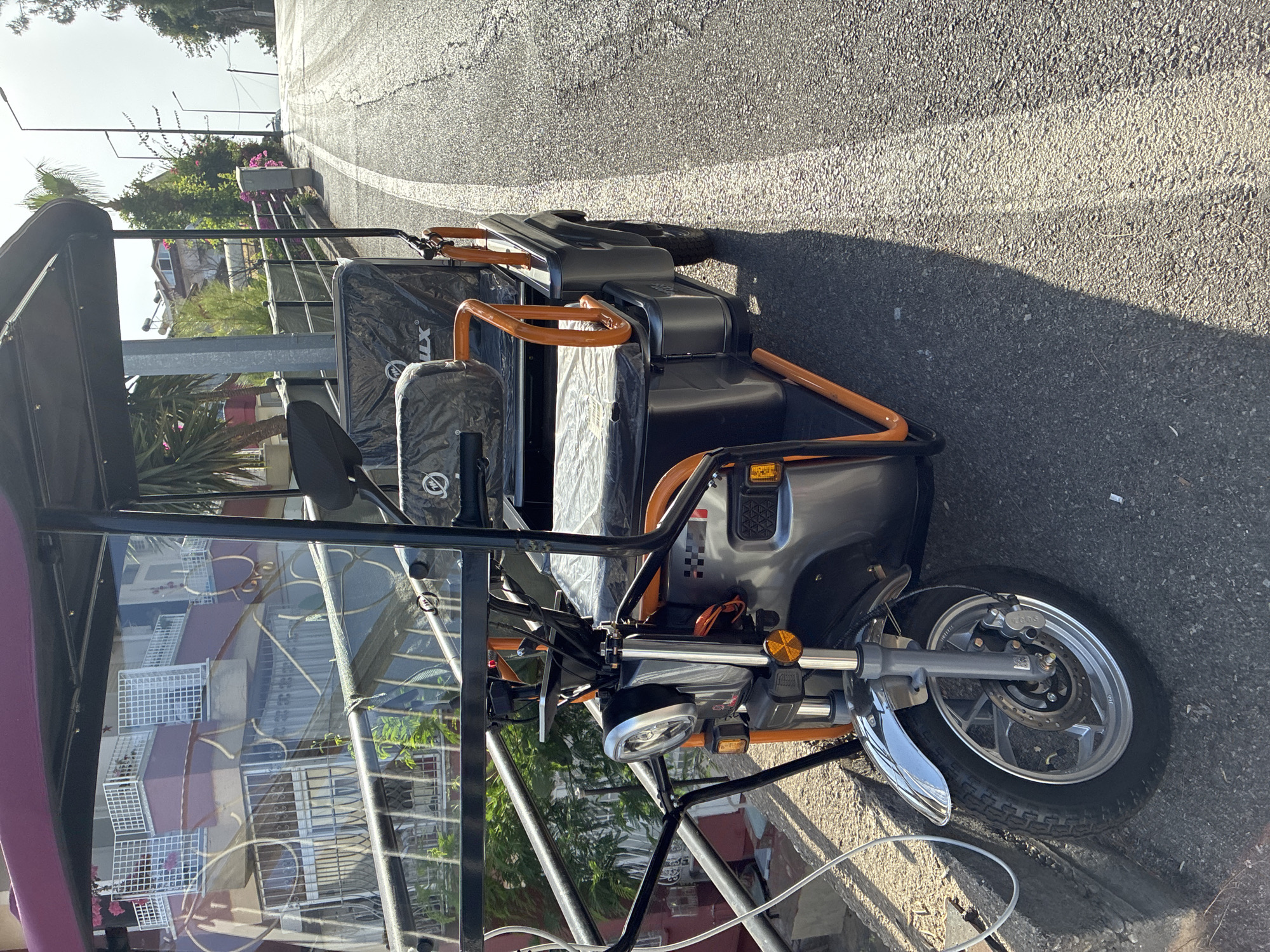 An electric three-wheeled vehicle, likely a "trike" or "tuk-tuk," is parked on a paved road in Alanya, Turkey, on an incline. The vehicle is dark gray with bright orange accents on its frame and handlebars. It has a covered seating area with clear plastic sides, a black canopy, and a single headlight. The vehicle appears to be stationary and there are no people visible in or around it. The road is lined with a curb and sidewalk on the left, with a metal railing and some greenery behind it. In the background, there are buildings with balconies and rooftops, characteristic of Mediterranean architecture, and streetlights are visible along the road. The sun is casting shadows, indicating it is daytime. The overall impression is one of a quiet, sunny street in a residential or tourist area.