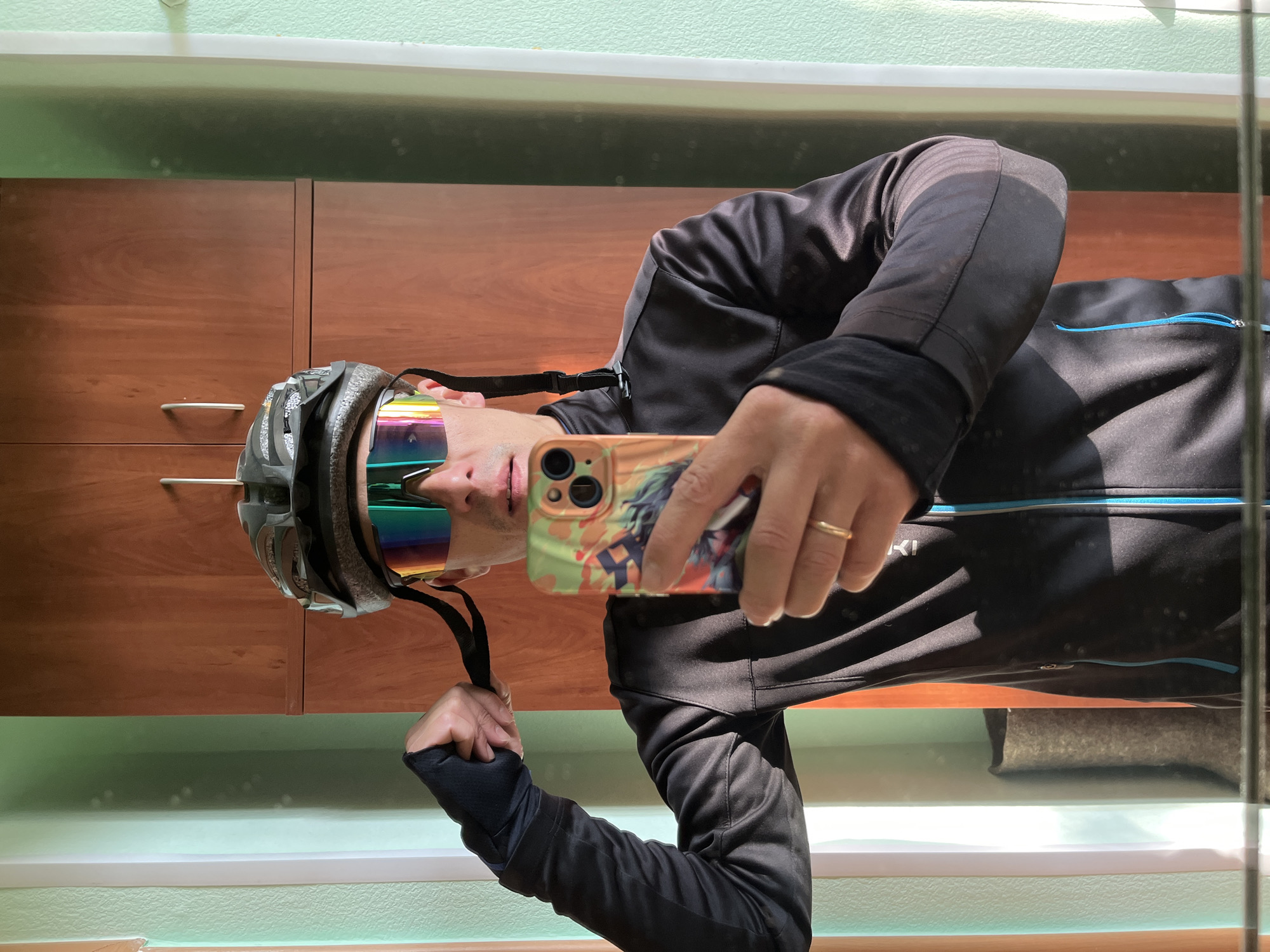 A man is taking a selfie in what appears to be an indoor environment. He is wearing a cycling helmet and iridescent sports sunglasses. He is wearing a black jacket with a blue zipper and holding a cell phone with a decorated case. The background includes a wooden cabinet with silver handles, a white windowsill, and a light green wall. The location cannot be determined from the image.