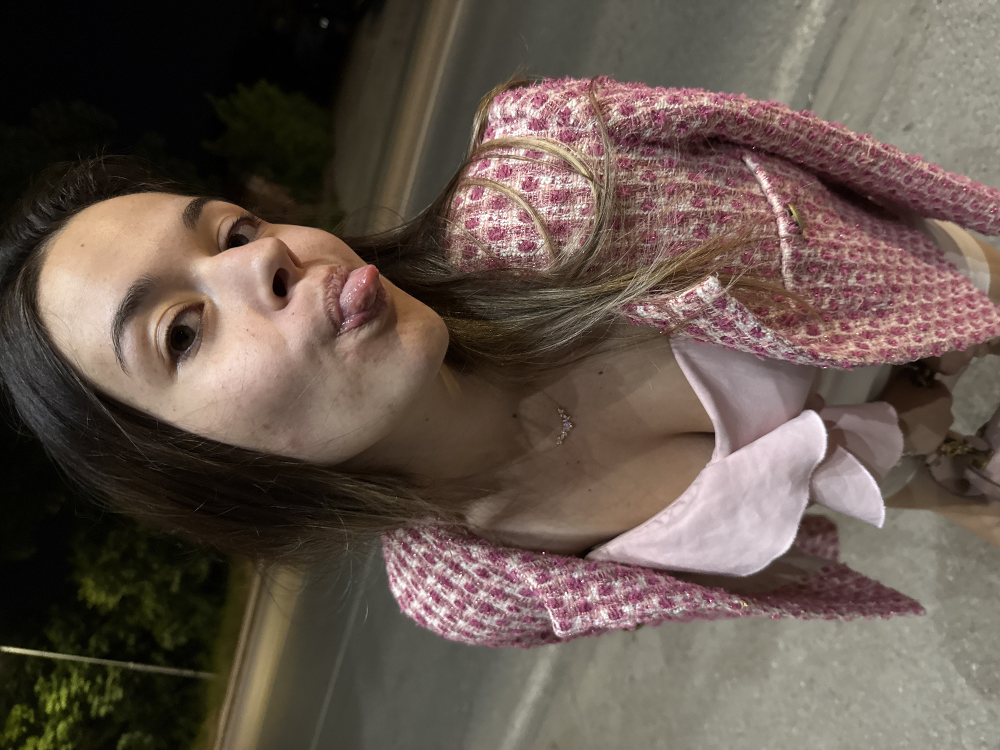 An outdoor nighttime portrait captured in Sofia, Bulgaria, features a young adult female posing on a paved surface, likely a street or sidewalk. The female, positioned centrally and angled slightly upwards towards the camera, has long dark brown hair and is looking directly at the viewer with a playful expression, her tongue extended. Her face exhibits minor visible skin imperfections. She is dressed in a textured, pink and white tweed-style jacket with shimmering threads, worn over a light pink camisole or top featuring ruffled fabric details around the décolletage. A delicate silver-colored necklace with small, possibly jeweled, clustered embellishments adorns her neck. The background is dark, revealing dimly lit green foliage from trees or bushes and a street light pole on the left side. The scene is illuminated by artificial light sources, consistent with a nighttime urban environment. No specific architectural landmarks or unique identifiers are visible to pinpoint an exact location within Sofia.