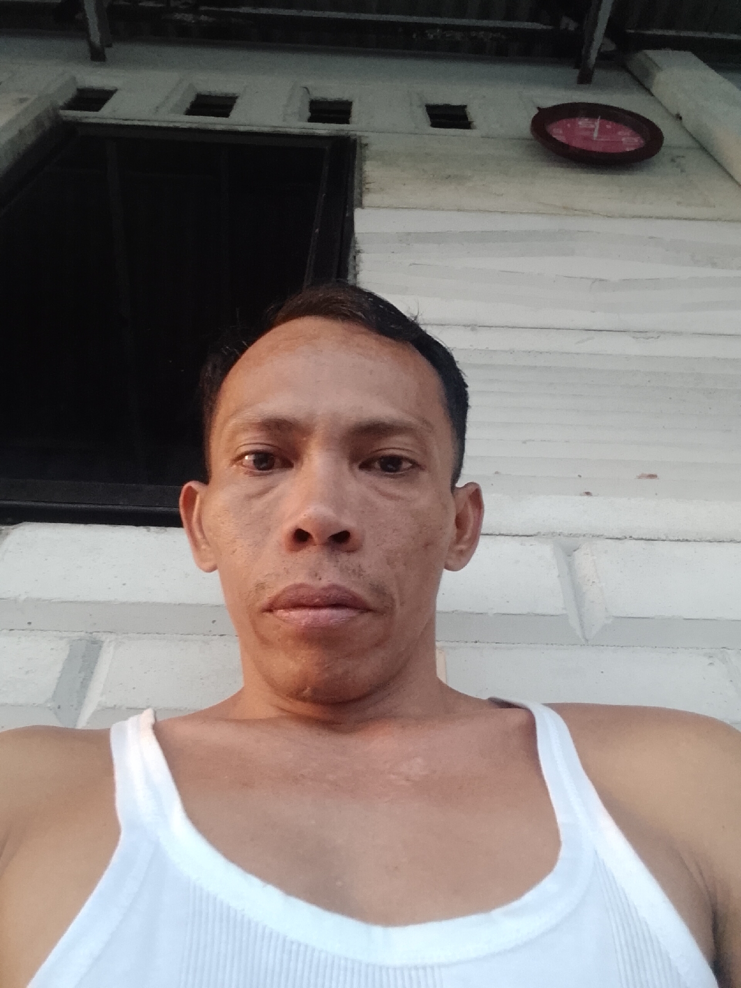 The image shows a man in a white tank top taking a selfie. He has dark hair and is in front of a white building with a window. There is a red clock on the wall to the right of the window. The image was taken in Bukit Ketok, Indonesia.