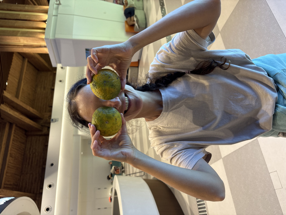 A woman stands in a modern, brightly lit indoor public space, likely an airport terminal or a large hotel lobby, situated in Tianya District, China. She occupies the center foreground, holding two round, green-yellow citrus-type fruits, each exhibiting dark surface blemishes, directly in front of her face, obscuring her eyes. She displays a wide smile with visible teeth. Her dark hair is secured in a long braid extending over her right shoulder. She wears a light-toned short-sleeved t-shirt and a light blue-green garment on her lower half. Her fingernails are manicured with light pink polish. The background features a high ceiling constructed with exposed dark wooden beams and lighter wooden panels, incorporating recessed circular lighting fixtures. The floor is tiled in a checkered pattern of alternating light grey and slightly darker grey squares. To the left, a curved white counter with a wooden trim is visible. To the right, a large white square pillar with vertical wooden slats on its upper section is illuminated, casting a warm glow. In the mid-ground, various lounge furniture pieces, including rounded green and orange seating pods, are present. One green pod is occupied by an individual lying prone. Additional blurred figures are visible in the background near the curved counter.