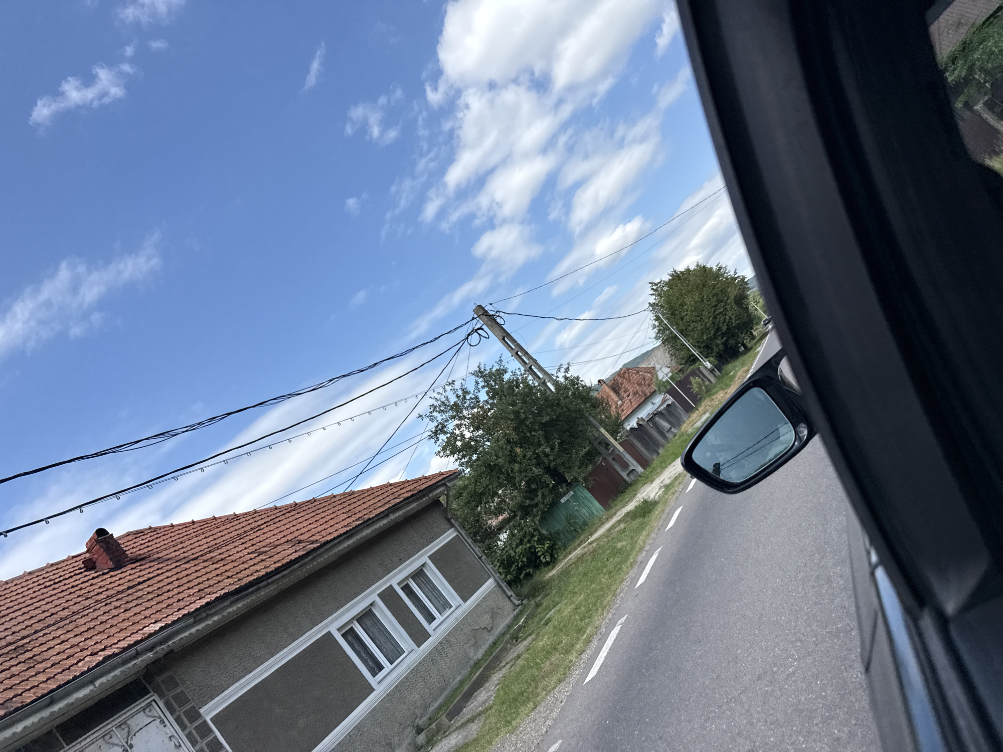 This image depicts a view from inside a car, looking out towards a street in Hârja, Romania. The scene is captured at an angle, with the car's interior visible in the foreground. A black car door and side mirror are prominent. The street is asphalt, with white markings. To the left, a building with a reddish-brown tiled roof and grey walls is visible, featuring several windows. A lush green tree stands to the right of this building. Further down the street, more houses with similar red roofs and wooden fences are present. Overhead, a bright blue sky is dotted with white, wispy clouds, and utility wires stretch across the frame, supported by a concrete pole. The overall impression is that of a rural or semi-rural setting on a clear day.