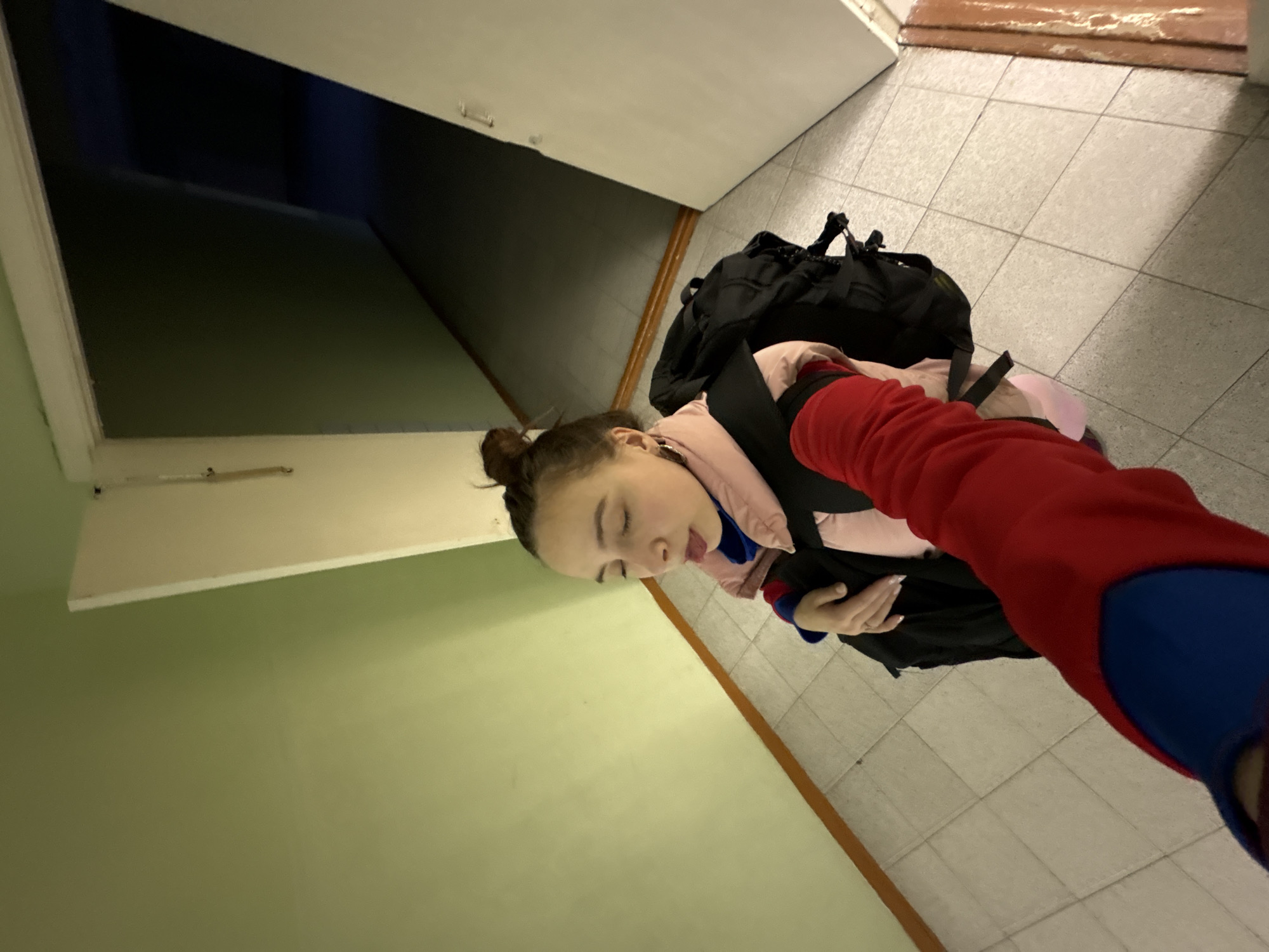 A woman stands in a hallway with tiled floors, wearing a pink puffer jacket, black backpack, and a red and blue hooded sweatshirt, with her hair in a bun. She is sticking out her tongue. The hallway has light green walls and a white ceiling with an open doorway leading to a dark area. The photograph is taken at an angle. The location is Berezniki, Russia.