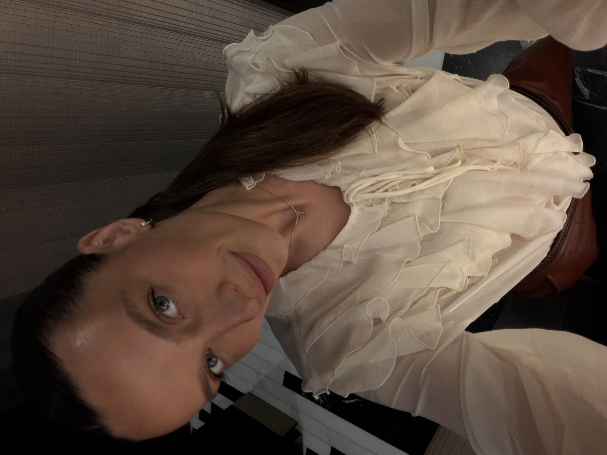In the city of Budva, Montenegro, a woman with dark brown hair pulled back into a ponytail takes a selfie. She is wearing a cream-colored blouse with ruffles and a v-neck that ties with a string. Her eyes are light blue, and she has a subtle smile on her lips. She is also wearing a thin silver necklace with a small pendant. The background is dimly lit, with vertical lines suggesting blinds or a textured wall, and a black and white patterned floor. Her lower half is obscured by her clothing, but a glimpse of a reddish-brown leather skirt or pants is visible at the bottom.