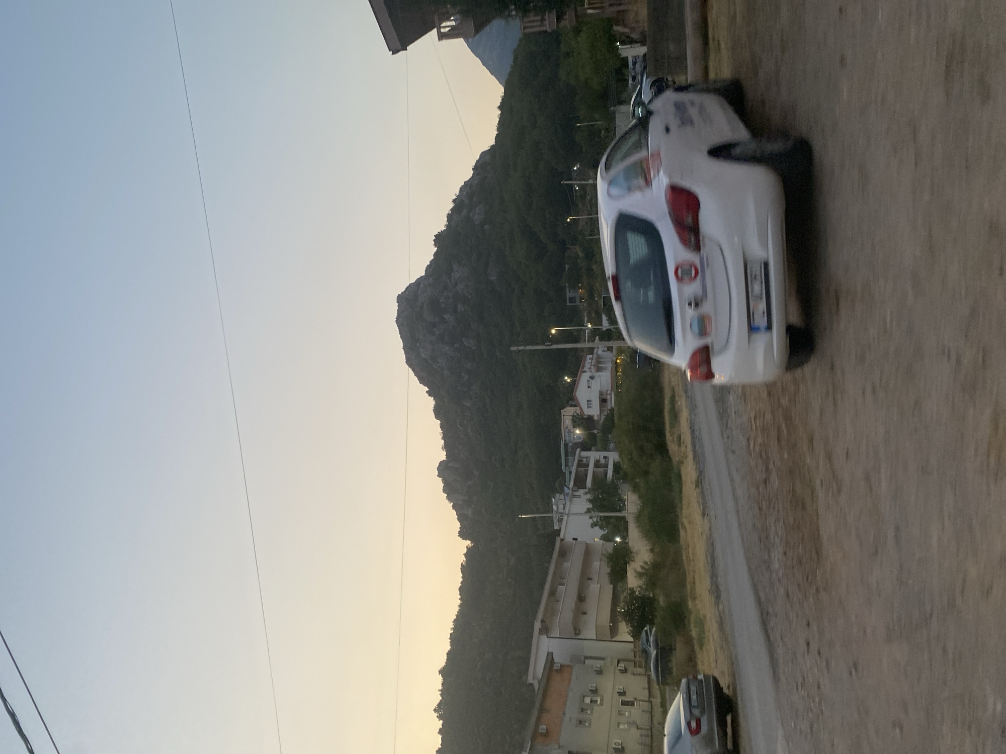 A white Opel Astra hatchback is parked on a dirt road in Sutomore, Montenegro, facing away from the viewer. The car is in motion, evidenced by the motion blur. The vehicle has a blue and white sticker on its rear bumper and a circular red and white sticker on the rear passenger door. The car is parked near a building on the right side of the frame. In the background, a lush green mountain range with rocky outcrops dominates the landscape. Several multi-story residential buildings with balconies and air conditioning units are visible to the left of the mountain, situated on a paved road. The sky is a soft gradient of pale yellow and light blue, suggesting it is either early morning or late evening. Power lines are visible crisscrossing the sky. The overall scene depicts a residential area in a mountainous coastal town.