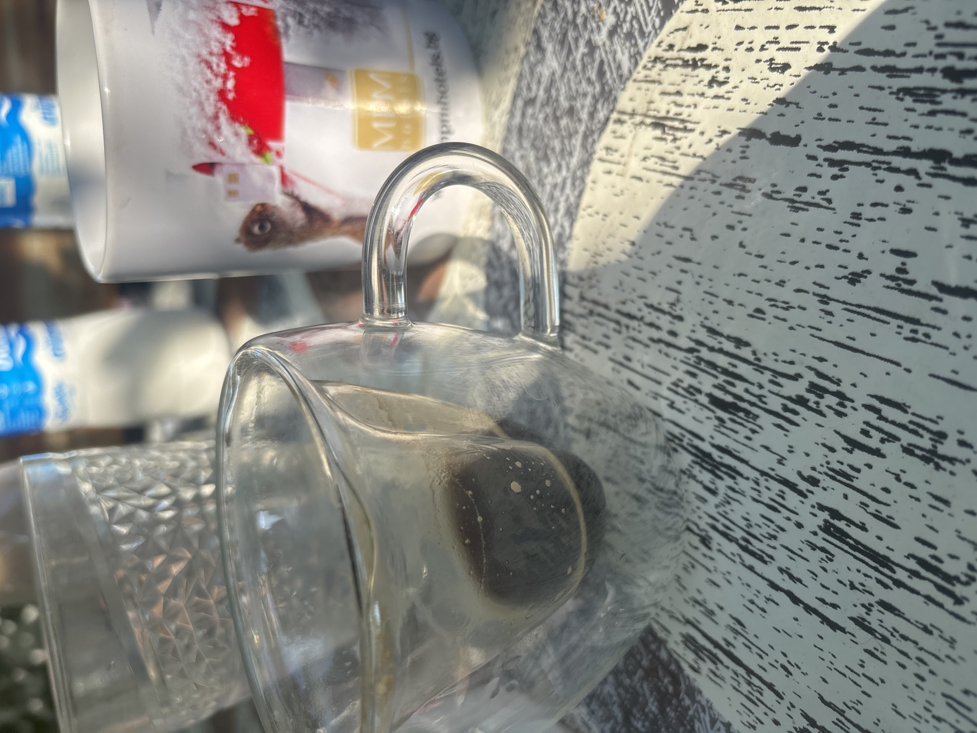 The image shows a close-up view of a glass cup with some remaining coffee at the bottom, placed on a table with a white and black patterned surface. In the background, there is a white mug with a picture of a squirrel and a snowy scene, and two out-of-focus bottles. The scene appears to be outdoors, with sunlight casting shadows on the table. There are no people visible in the image. The location is Trudovets, Bulgaria.
