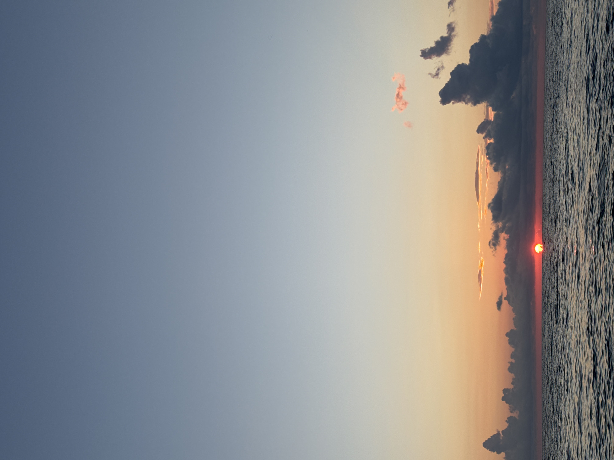 The image depicts a tranquil seascape at dusk, likely near Batumi, Georgia. The sky transitions from a pale blue at the top to warm orange and pink hues near the horizon. A vibrant red sun, partially obscured by dark, dramatic clouds, is setting over the dark, rippling water of the sea. The clouds are scattered, with some appearing dark and heavy, while others are lighter and wispy, tinged with the colors of the sunset. The overall scene conveys a sense of peace and natural beauty.