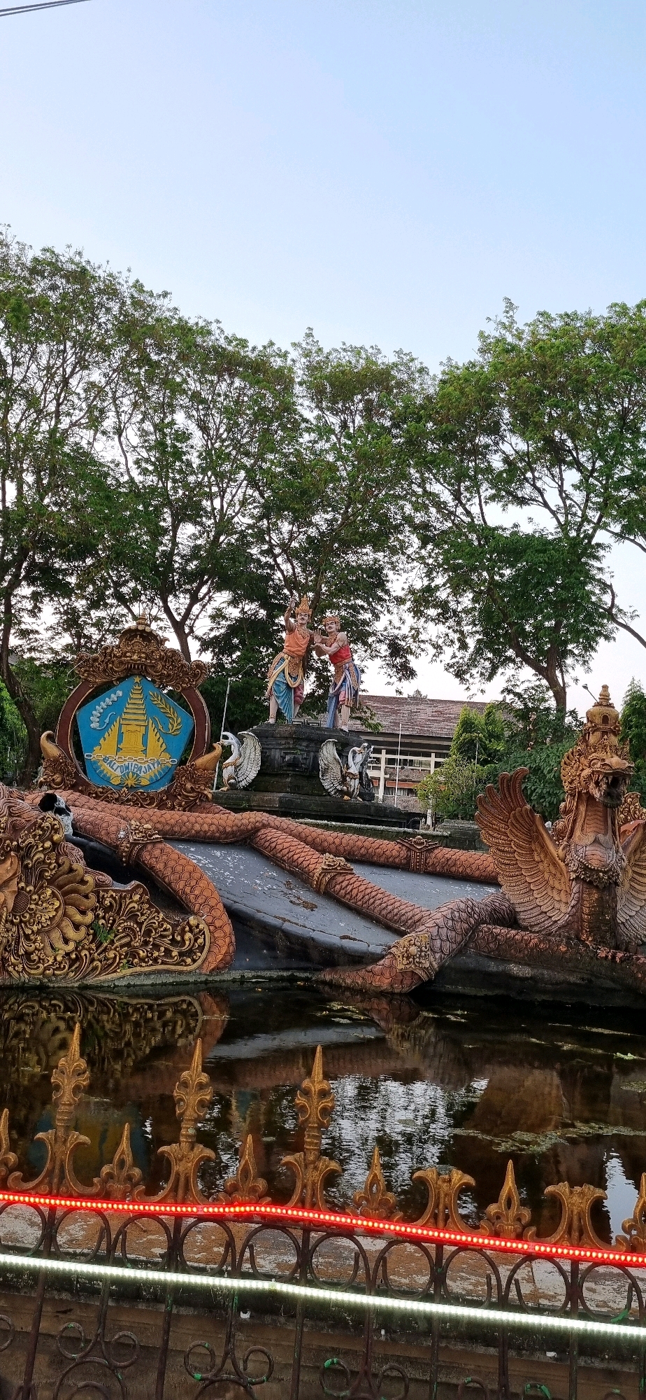 The image shows a decorative fountain in Denpasar, Indonesia, featuring statues and intricate carvings. In the background, lush green trees line the horizon under a clear sky. In the foreground, a pond reflects the surrounding structures and sky. The fountain itself is adorned with a crest bearing the inscription "BALI DWIPA JAYA," and two statues of figures in traditional attire are positioned above the crest. To the right of the central figures, a mythical bird-like creature with outstretched wings is sculpted. The pond is bordered by a decorative metal fence with red and white LED lights. The overall scene depicts a cultural landmark in Denpasar, likely a public space or a monument.