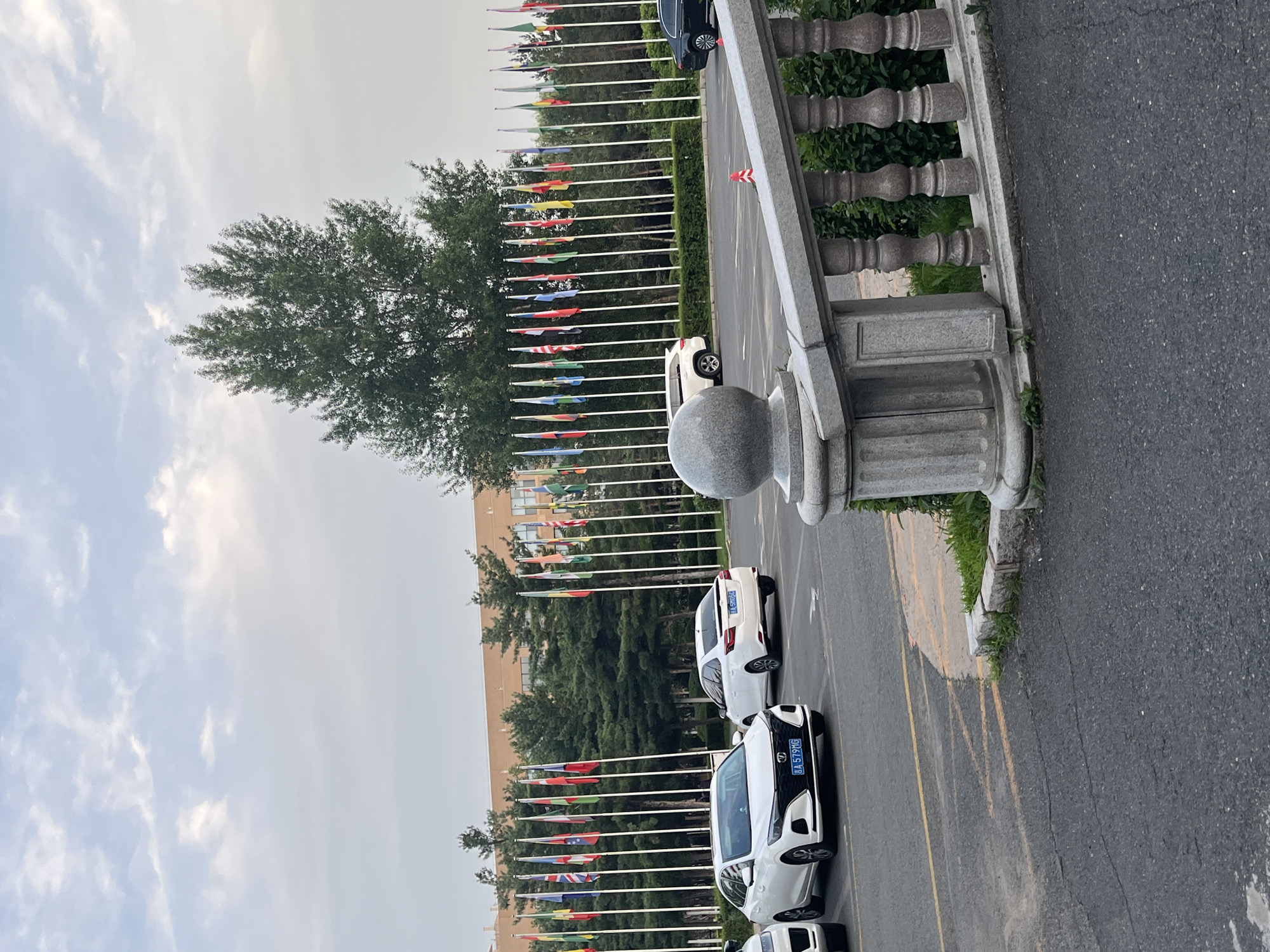The image depicts an outdoor scene, likely a public space or an institutional campus, featuring a row of national flags displayed on poles. Several white sedans and a white SUV are parked in what appears to be a parking lot in front of the flags. A building with an orange facade is visible in the background, behind the flags and a line of trees. In the foreground, a stone balustrade with ornate pillars lines a raised area. The sky is overcast with scattered clouds. The scene is static, with no visible human activity. The presence of numerous national flags suggests a diplomatic or international setting. A digital license plate on one of the white sedans reads "A 579 MG" and another reads "A 579MG". Based on visual cues, the location is identified as the Jilin University of Finance and Economics in Changchun City, China.