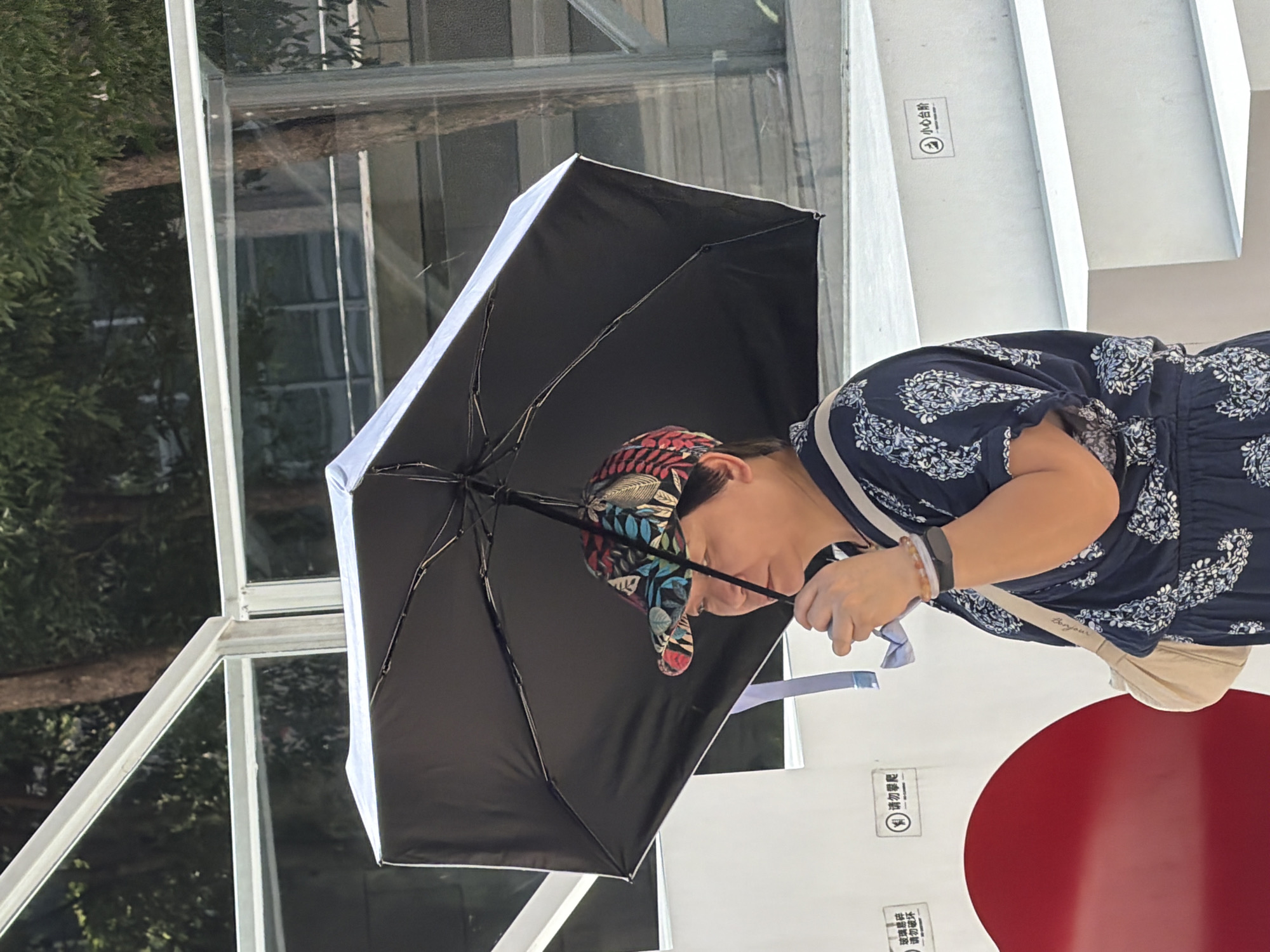 In Yuzhong District, China, a person is holding a black umbrella open overhead, shielding themselves from the sun. They are wearing a dark blue dress with a white floral pattern and a colorful cap. A beige crossbody bag is slung over their shoulder. The person is standing near a white structure with a large red circular element, and a set of white stairs is visible behind them. Signs with Chinese characters are affixed to the white structure and the stairs. The background shows trees and a glass enclosure.