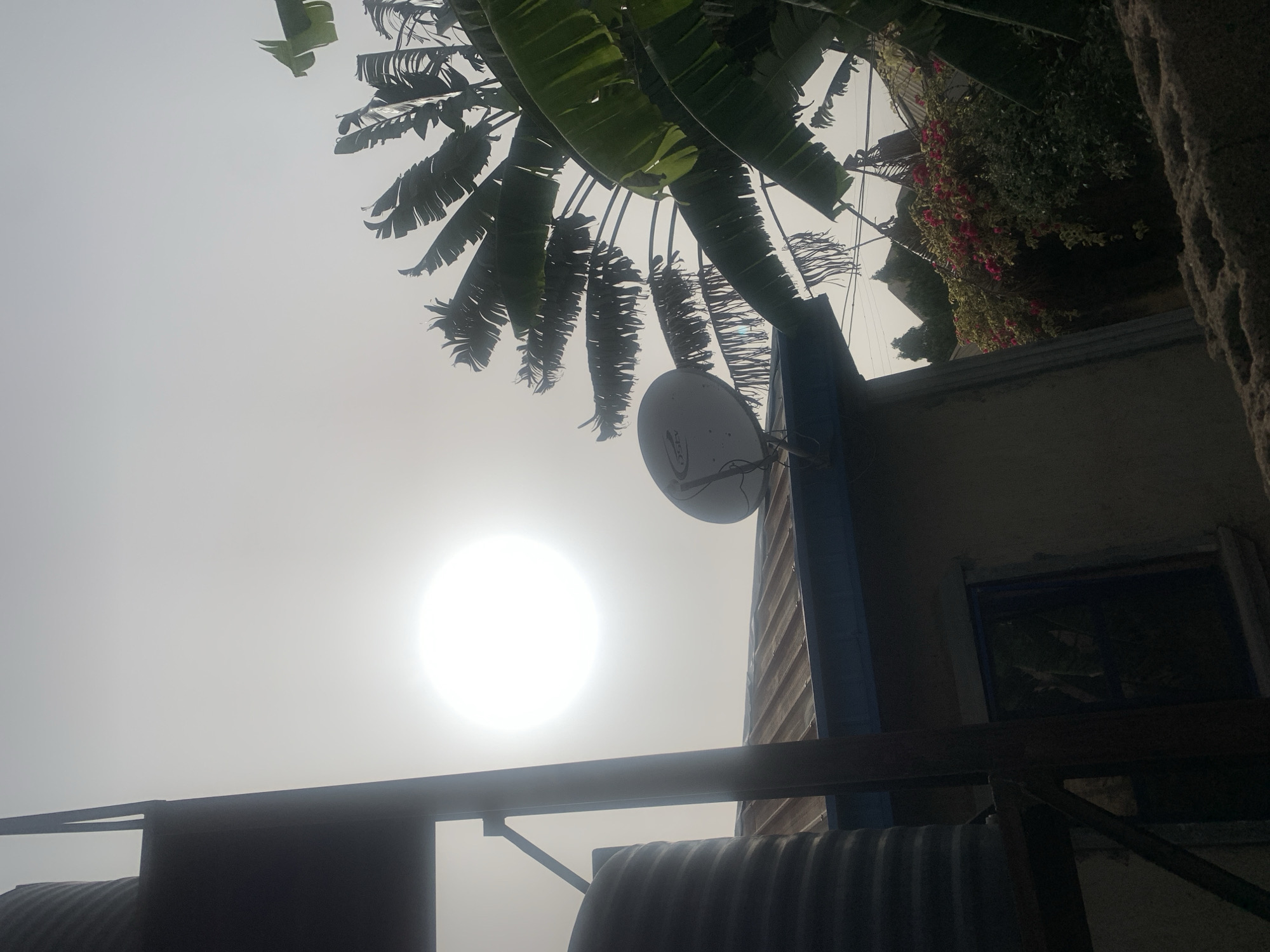 The image shows an overcast sky in Barakin Akawo, Nigeria, with the sun as a bright, diffused orb. In the foreground, there's a partially obscured view of a corrugated metal structure with dark beams. A satellite dish is mounted on a blue support structure attached to a building with a window. Overhanging the building is a banana tree and some flowering foliage. The overall scene is viewed from a low angle.