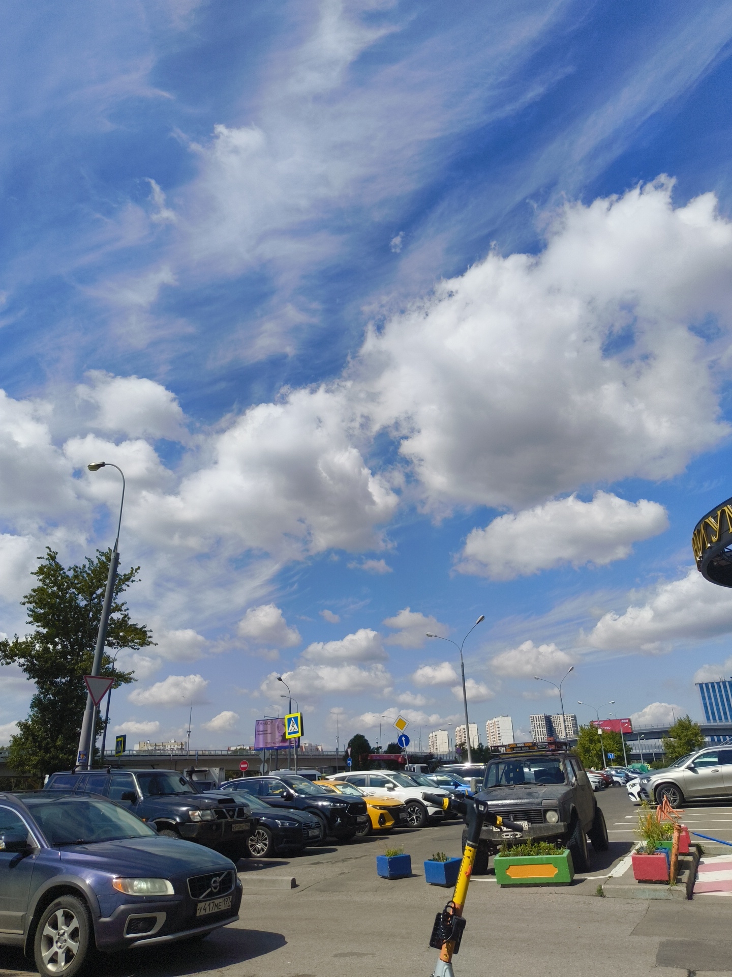 The image depicts an outdoor parking lot under a partly cloudy sky. In the foreground, a dark blue Volvo SUV is parked, facing right. Behind it, a line of cars including a black SUV, a black Audi, and a yellow car are parked. A dark-colored Niva-style vehicle with a roof rack and some greenery in a planter box is also visible. The background shows a cityscape with buildings and trees, and streetlights. A decorative circular structure with golden lettering is visible on the right side of the image. The overall scene suggests a commercial or public parking area.