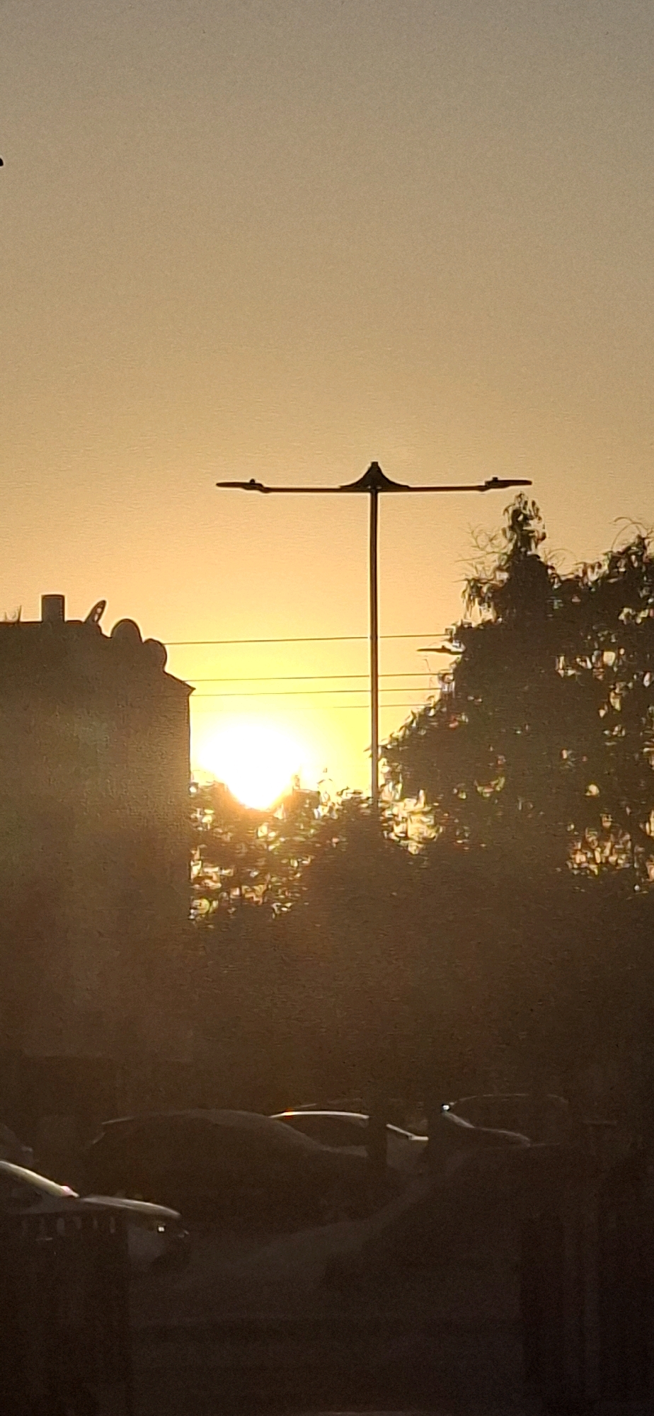 The image depicts a sunset scene in the city of Yerocham, Israel. In the foreground, silhouettes of parked cars are visible, appearing as dark shapes against the brighter background. Further back, a building with an ornate roofline is partially obscured by the glare of the setting sun, which is positioned in the center of the frame. A tall street lamp with a horizontal fixture stands prominently against the sky, with its base extending down towards the middle ground. Trees with dense foliage are seen on the right side of the image, their forms softened by the sunset light and haze. The sky is dominated by a warm, orange-yellow gradient, characteristic of a sunset. The overall scene captures a moment of tranquil urban twilight.