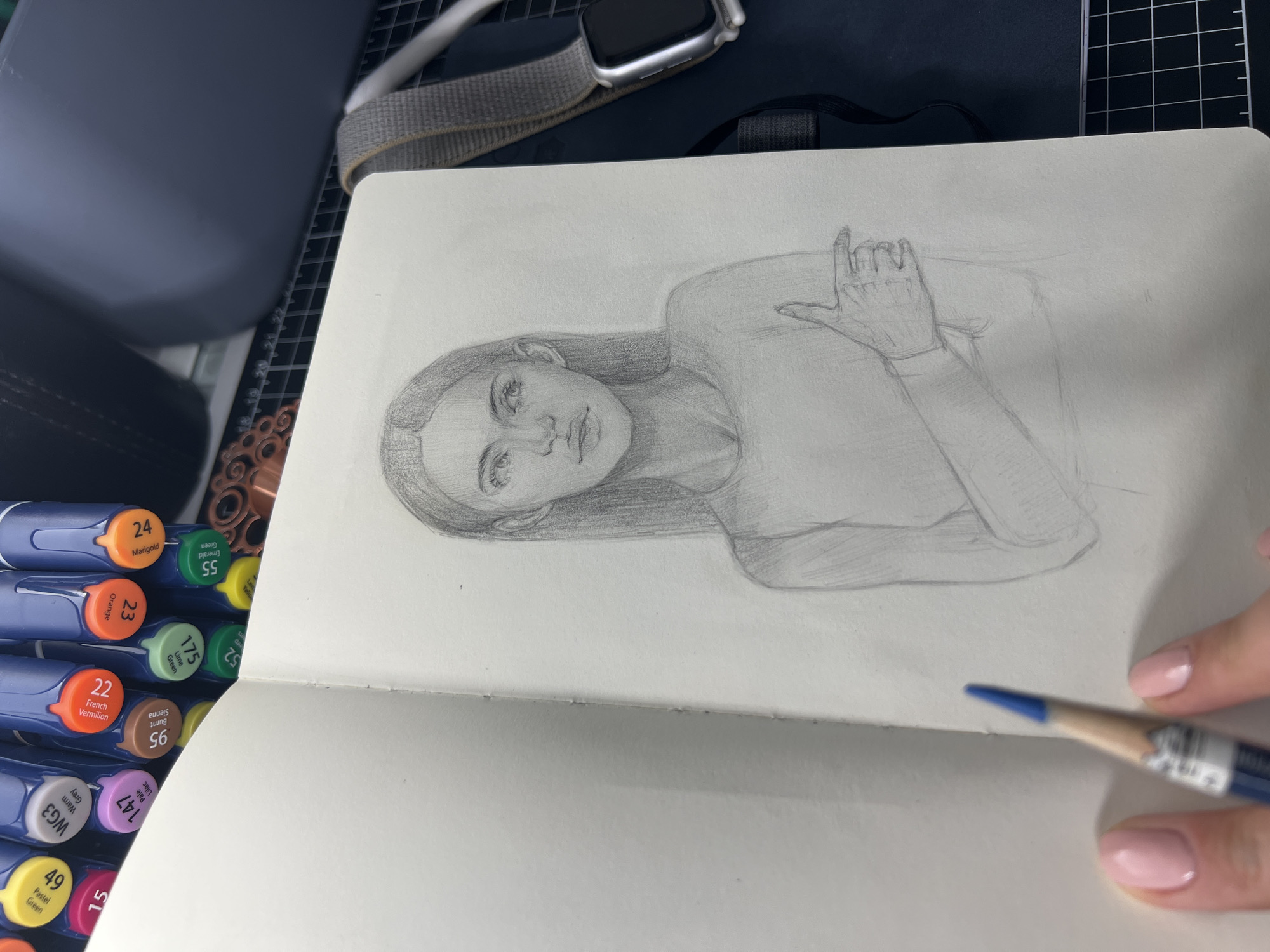 The image shows a pencil sketch of a young woman in a notebook. The sketch is of her upper body, with her head tilted to the left and her right arm bent across her chest, pointing to the left. Her hair is long and dark, falling around her shoulders. She is wearing a long-sleeved top. The notebook is open, revealing a blank page on the left and the sketch on the right. To the left of the notebook, a hand with pink painted fingernails holds a blue pencil, poised to draw or erase. In the background, a set of colorful markers are visible, along with a dark surface with a grid pattern, a dark grey object, and a smartwatch with a grey band. The scene appears to be an artist's workspace, likely indoors.