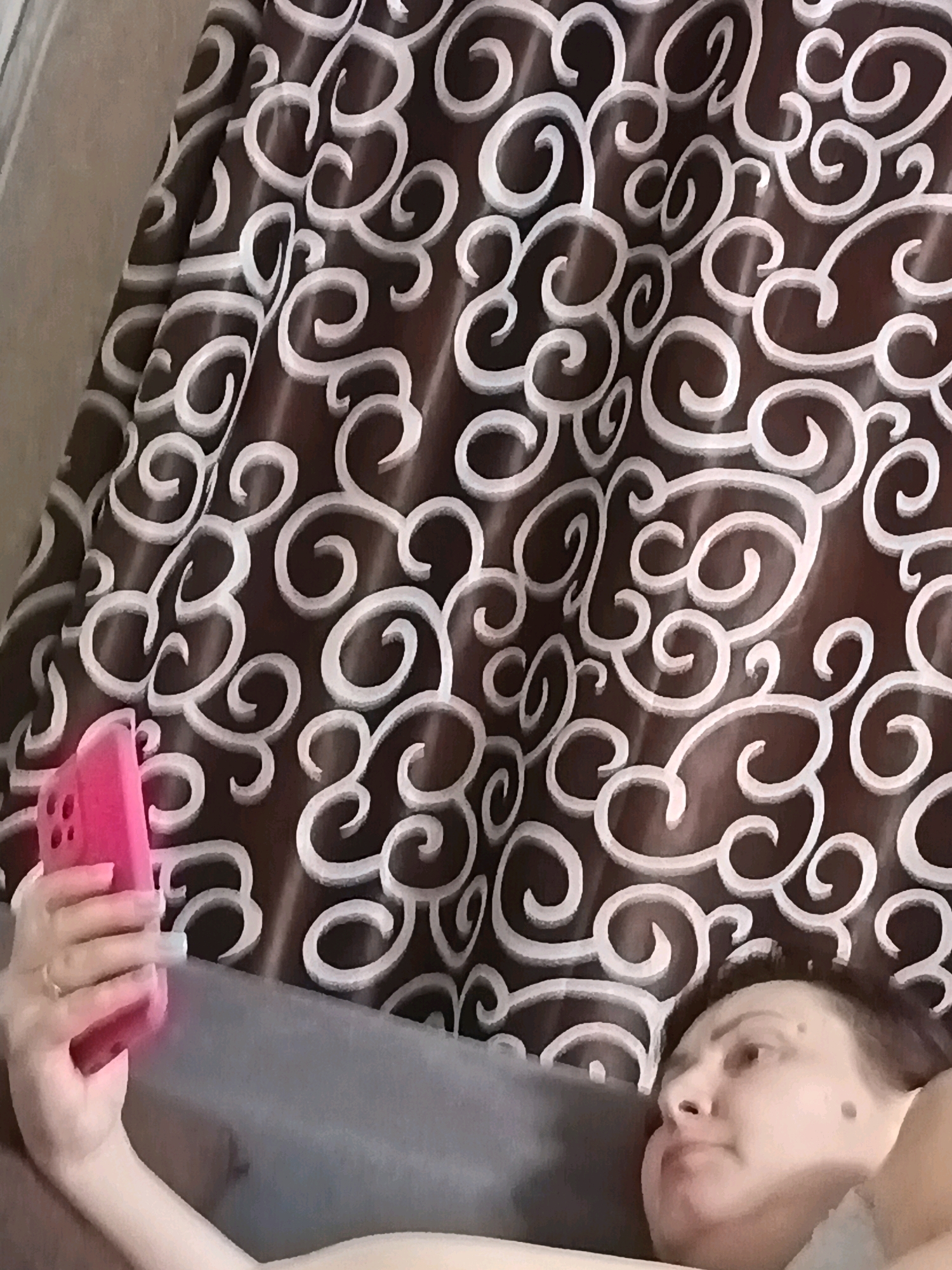 A woman is taking a selfie with her pink smartphone. She is lying down on a grey surface, possibly a sofa or bed, with a brown and white patterned curtain in the background. The woman has dark hair and two moles on her right cheek. Her left hand is holding the phone, and she is wearing a ring on her left ring finger. The lighting appears to be indoor, and the overall mood is casual and relaxed. The image does not provide any specific information about its location in Astrakhan, Russia.