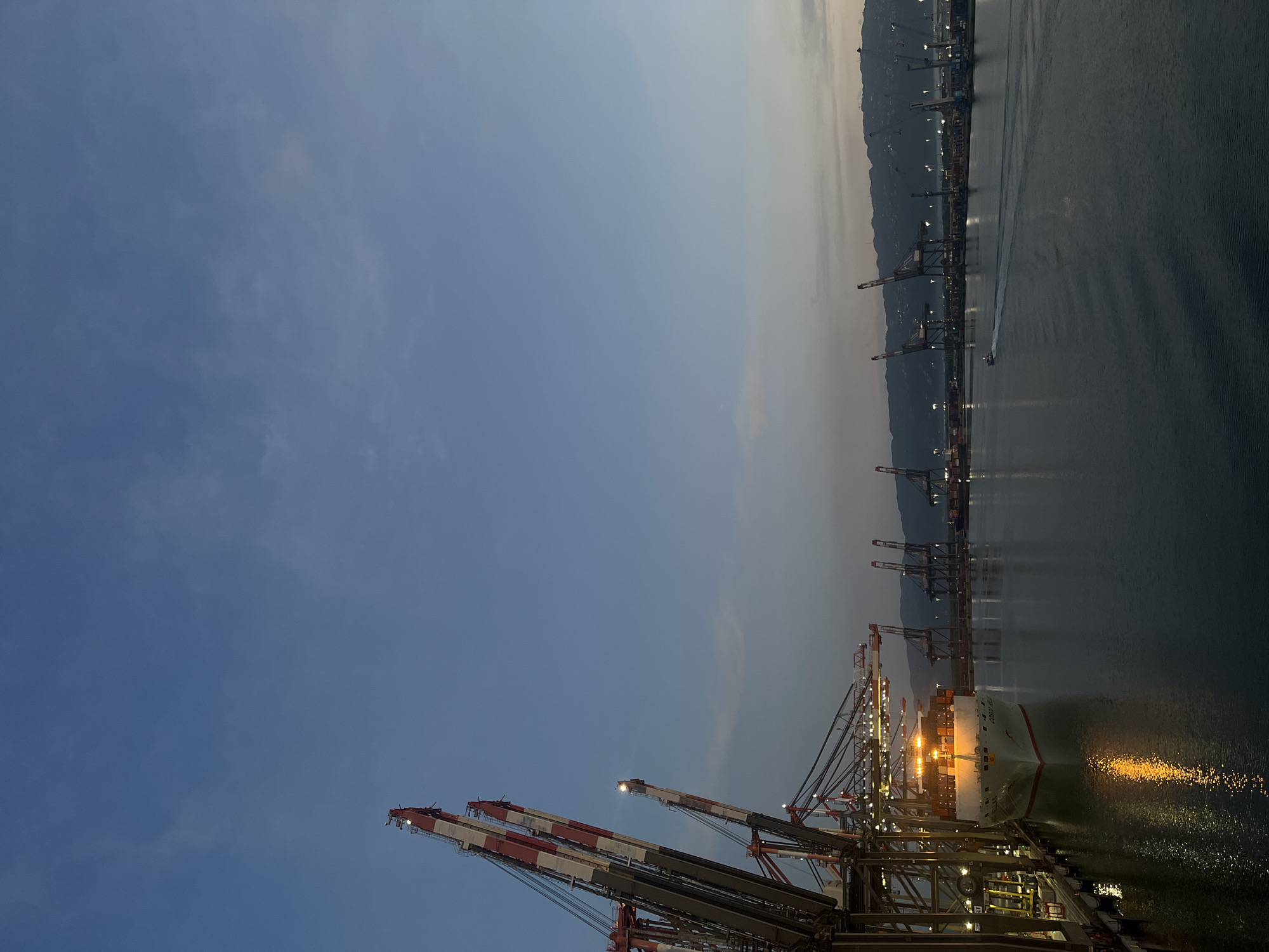 The image depicts a port at dusk. A large cargo ship is docked, illuminated by artificial lights. Cranes tower above the ship, also illuminated. The water reflects the lights, creating shimmering patterns. In the background, the coastline is visible, marked by a series of lights, possibly indicating a city or industrial area. A small boat is visible on the water, leaving a wake. The sky is a gradient of blue and grey, suggesting the transition from day to night. The image's orientation is vertical.
