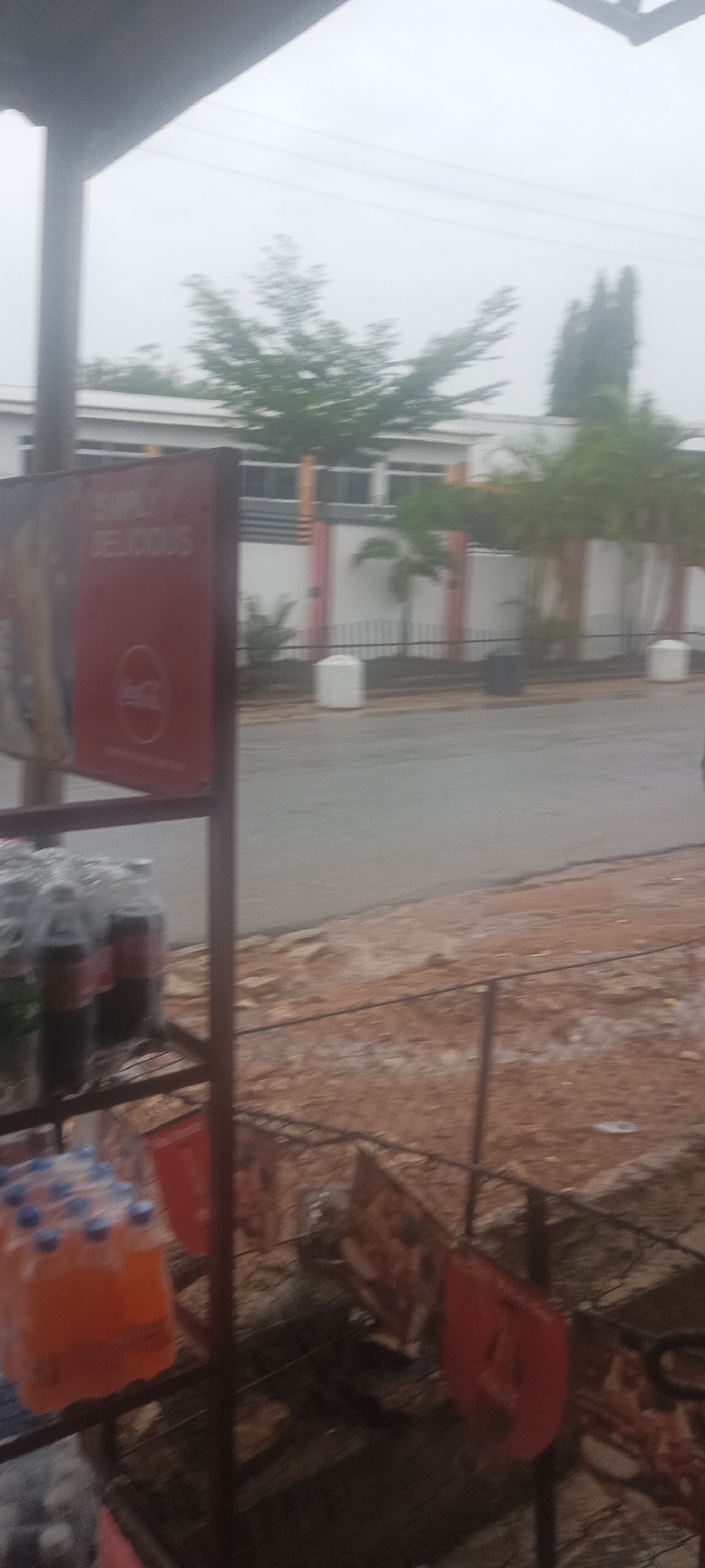 The image shows a street scene in Jos, Nigeria, viewed from a storefront. A metal rack displays bottled drinks, including Coca-Cola and Fanta. Signage for Coca-Cola is attached to the rack. Across the street, a white building with a grey roof and orange vertical accents along its fence is visible. Palm trees and other greenery line the fence. The sky is overcast, and the street appears wet. The foreground area near the storefront is unpaved and has exposed reddish soil.