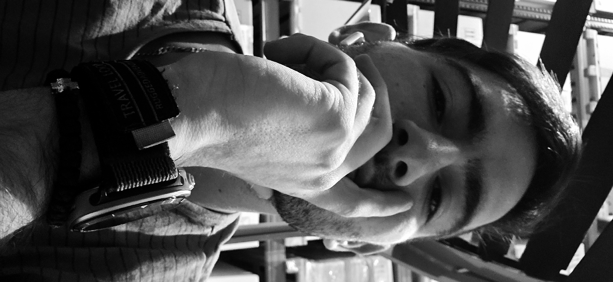 A close-up, monochrome portrait shows a man with dark hair, stubble, and partially closed eyes, positioned from the upper left to the lower right of the frame. He is looking downwards with his face tilted. His right hand is raised and held to his face with his fingers partially covering his mouth and chin. A watch with a thick band is visible on his wrist. The watch band has the words "TRAVEL FOR" and "RUGGED" printed on it. The background consists of blurry, indistinct elements including what appears to be shelves with items on them and structural supports. The photograph does not offer information about the location of Plovdiv, Bulgaria, or suggest any connection to that city.