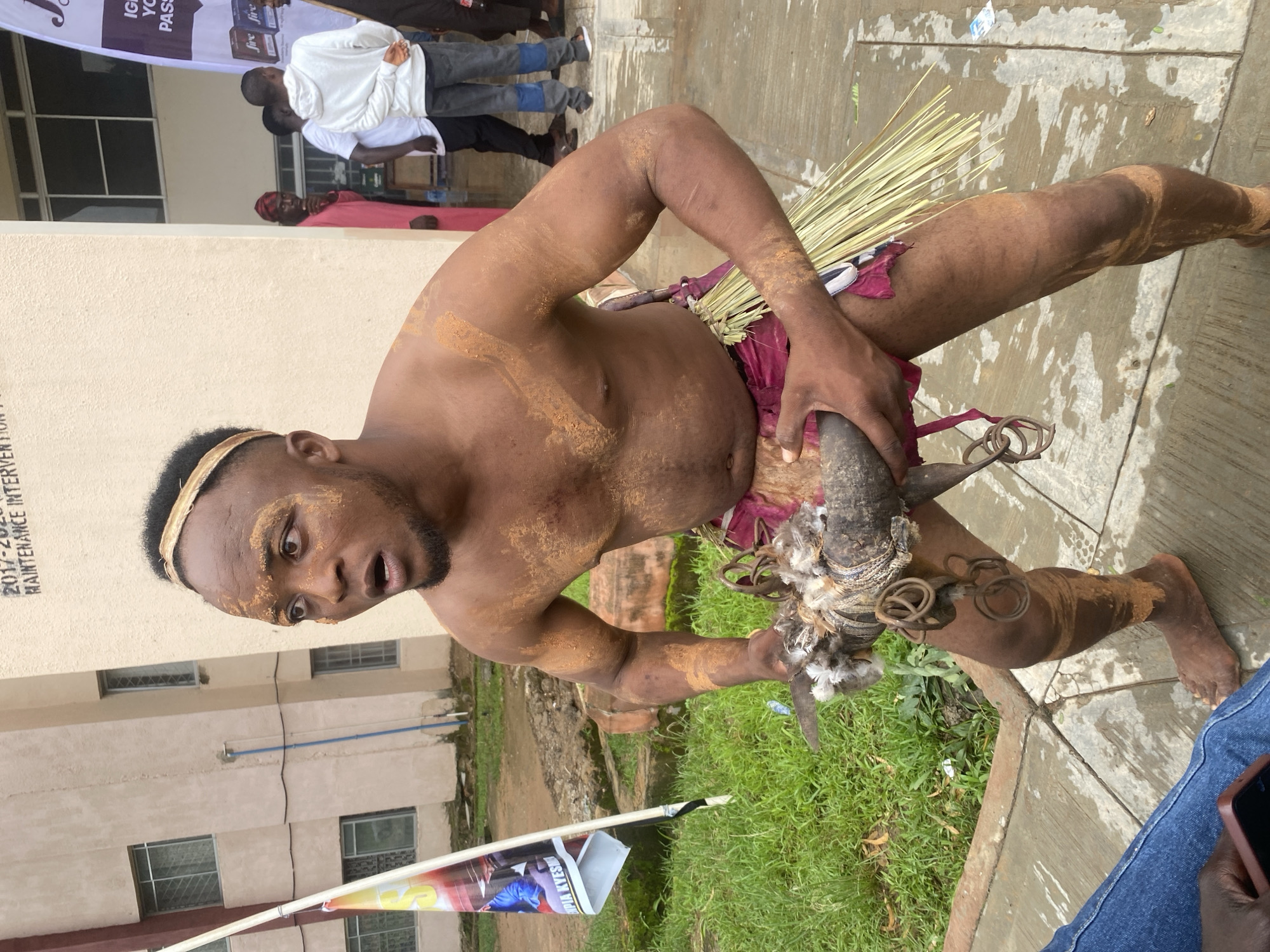 The image captures a man with his upper body exposed, adorned with what appears to be ochre or mud, standing on a concrete walkway. He is wearing a burgundy-colored loincloth tied with a straw-like material. His face is turned to the left, with his mouth open in a surprised or exclamatory expression. He is holding a dark, cylindrical object with feathers and metal rings attached in both hands, positioned in front of his torso. The background features a light-colored, textured building with visible windows and a blue pipe running vertically along the wall. To the left of the man, a white pole with a flag is partially visible, displaying colorful graphics. The ground in front of the walkway is covered with green grass. In the background, several other individuals are visible standing near the building entrance. One person wears a pink garment, another a white hoodie, and others are dressed casually. A banner with text and logos is also visible in the background. The overall scene suggests an outdoor cultural event or performance. The location is identified as Rafin Par, Nigeria.