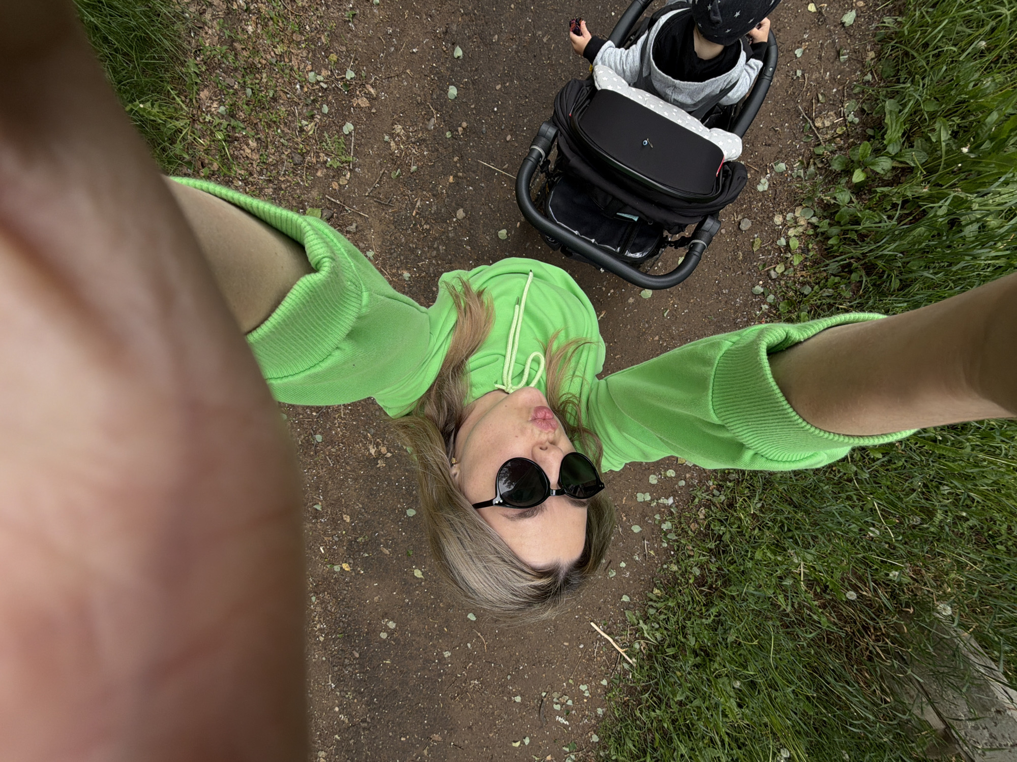 An overhead, high-angle selfie captured in Chaikovsky, Russia, features a woman in a bright green hoodie and a baby in a stroller on a dirt path. The woman, with blonde hair and sunglasses on her face, is looking up at the camera, making a puckered or kissing face. Her arms and part of her body are visible as she holds the camera from above.
Below her, to the right, a baby or toddler is seated in a black and grey stroller. The baby is wearing a grey and black outfit and a black hat with white stars, and appears to be holding a small item. The stroller is black with grey accents.
The setting is outdoors, likely a park or walking trail. The ground beneath them is a dirt path mixed with sparse green grass and small leaves. To the right side of the frame, a patch of thicker green grass is visible. The lighting suggests an overcast or cloudy day, as there are no distinct shadows. The overall scene conveys a casual, playful moment during an outdoor stroll. No visible text is present in the image.