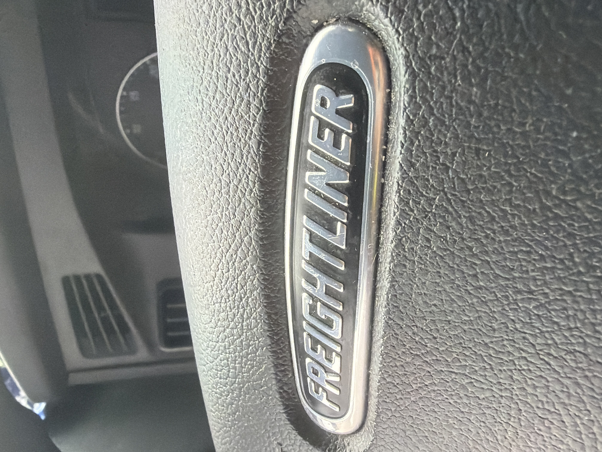 A close-up, eye-level, horizontal shot shows the "FREIGHTLINER" logo on the steering wheel of a truck. The logo is a silver oval with raised, capitalized lettering. The steering wheel is made of black, textured leather. In the background, out of focus, are parts of the truck's dashboard, including air vents and a circular gauge. The image appears to have been taken during the daytime, with bright lighting conditions. The location of this image is not specified in the provided data.