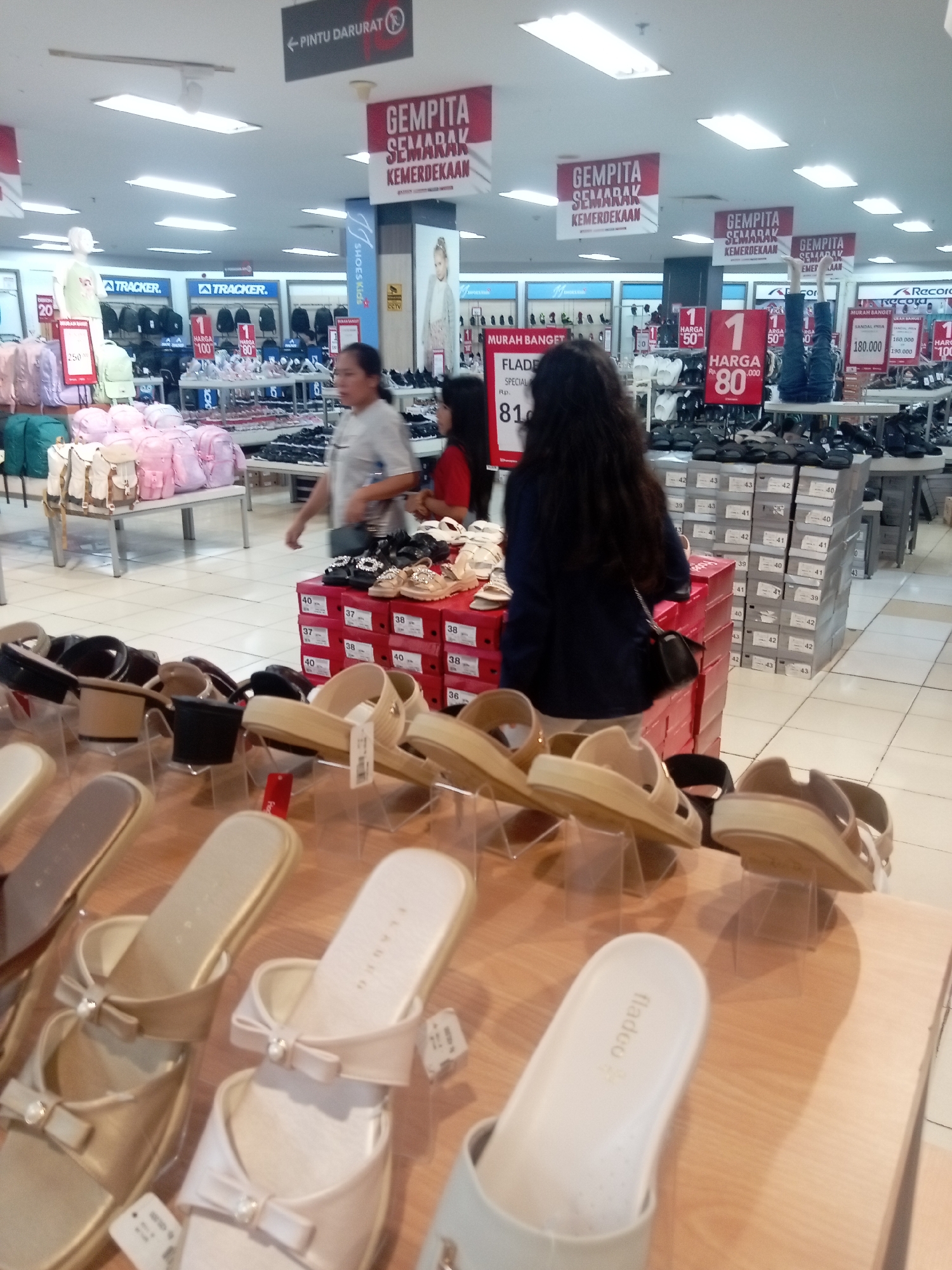 The image shows the interior of a shoe store, likely located in Batam, Indonesia, as indicated by the "GEMPITA SEMARAK KEMERDEKAAN" banners, which are Indonesian for "Festive Independence Buzz." The store is well-lit with fluorescent ceiling lights. In the foreground, several pairs of women's sandals are displayed on wooden shelves. These sandals are in various neutral colors like beige and white, some featuring decorative bows and pearls.
Further back, a woman in a gray shirt is walking, and another woman in a red shirt is visible behind a display of shoes. Behind them, stacks of shoe boxes are neatly arranged, with some boxes displaying sizes like 40, 42, 43, and 39. Various brands of shoes and bags are visible on display racks and mannequins in the background, including "TRACKER" and "Record." Promotional banners indicating discounts and prices, such as "HARGA Rp. 80.000" (Price Rp. 80,000), are hung from the ceiling and on display stands. The floor is tiled with a light-colored pattern. The overall environment suggests a busy retail setting with a sale event in progress.
