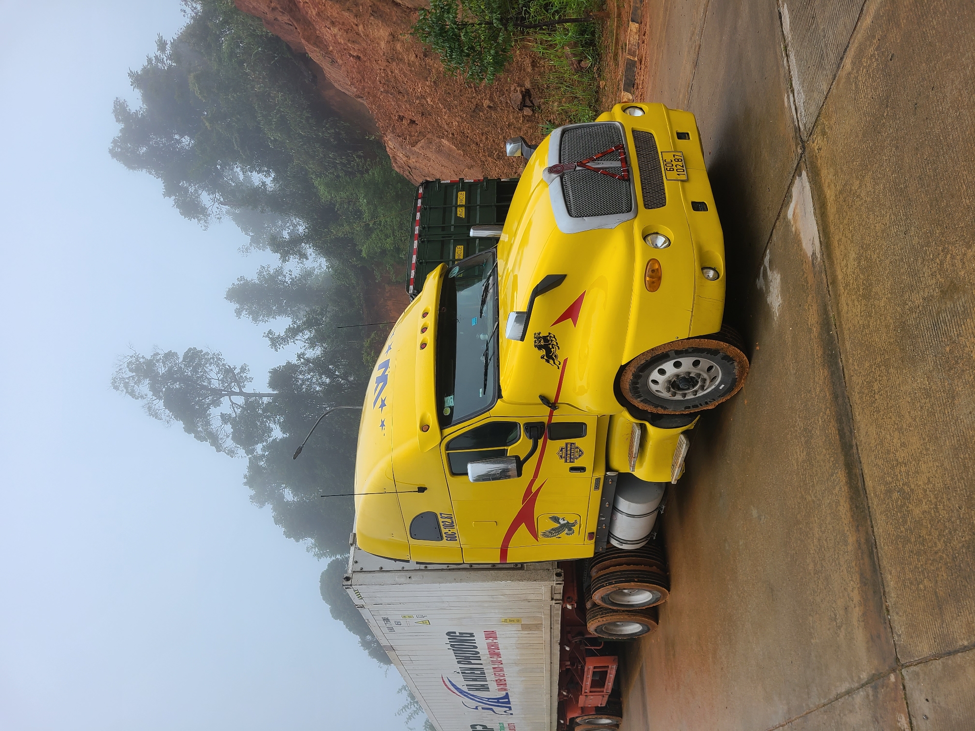 A bright yellow semi-truck and trailer is parked on a concrete surface. The truck's cab has red and black decals, including a red lightning bolt and an eagle graphic. The trailer is white with blue text that reads "HÀ HIỀN PHƯƠNG" and other smaller text in Vietnamese and English, including "VIET NAM - CAMPUCHIA - CHINA." The truck has a license plate with the numbers "60C 102.87." In the background, there is a steep, reddish-brown hillside with trees and foliage, and a dense fog obscures the upper portions of the trees and the sky. Another dark green truck is partially visible behind the yellow semi-truck. The concrete surface is wet and stained with reddish-brown mud, indicating recent rainfall or a muddy environment. The overall scene suggests the truck is on a road or a rest stop in a rural or mountainous area with poor visibility due to fog. The location is Xayden, Laos.