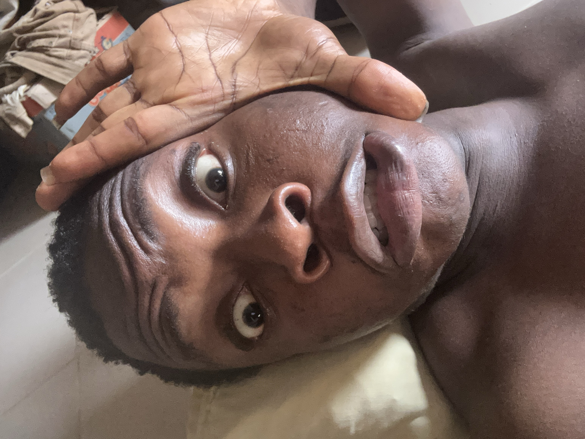 A close-up shot of a person of color lying down with their hand on their forehead. The person's eyes are open, looking up at the camera. Their lips are slightly parted. The background is blurred and out of focus. The scene is indoors.