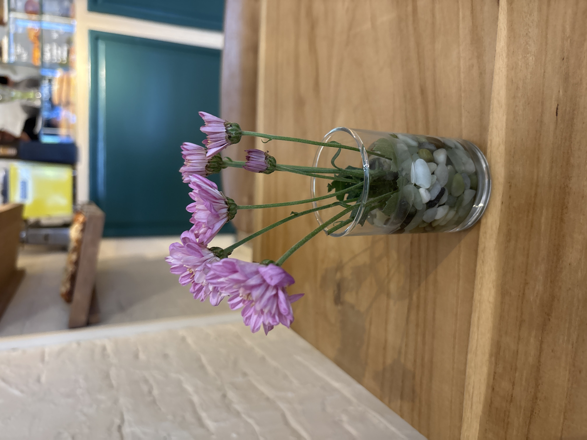A glass vase filled with green and white pebbles is laying on its side on a wooden surface. The vase contains several purple flowers. The setting is indoors. The location is likely Kuta Selatan, Indonesia.