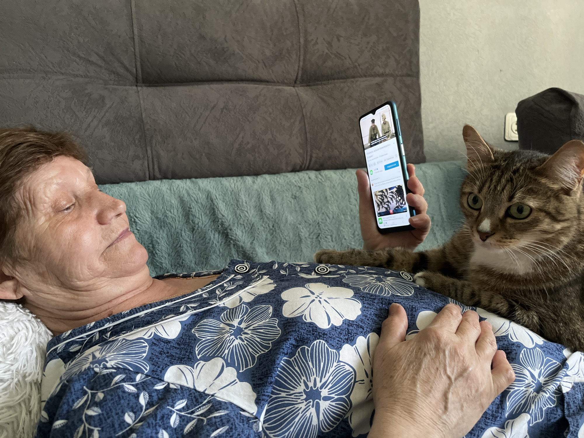 A woman is lying in bed, holding a cell phone in her right hand, with a cat sitting next to her. The woman is lying on her left side, wearing a blue and white floral-patterned top. She is looking at the cell phone screen. The cat, with brown and white fur, is sitting upright and looking forward. The cell phone screen shows a video of two men in military uniforms. The background includes a gray headboard and a light blue blanket. The location is unknown and cannot be determined from the image.