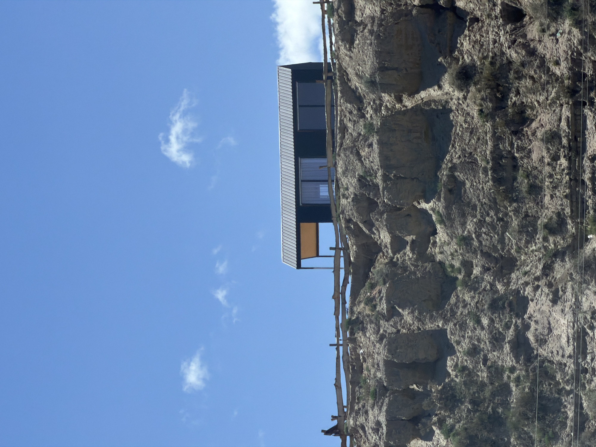 A modern, dark-colored dwelling with a silver, corrugated metal roof and large windows sits atop a rugged, dry hillside under a bright blue sky with scattered white clouds. A rustic wooden fence runs along the edge of the hill, separating the dwelling from the steep drop. The hillside is characterized by exposed rock formations and sparse vegetation. A lone figure is visible on the left side of the fence, appearing to be standing and looking towards the dwelling. The scene is captured in daylight, suggesting an outdoor setting in a remote or elevated area.