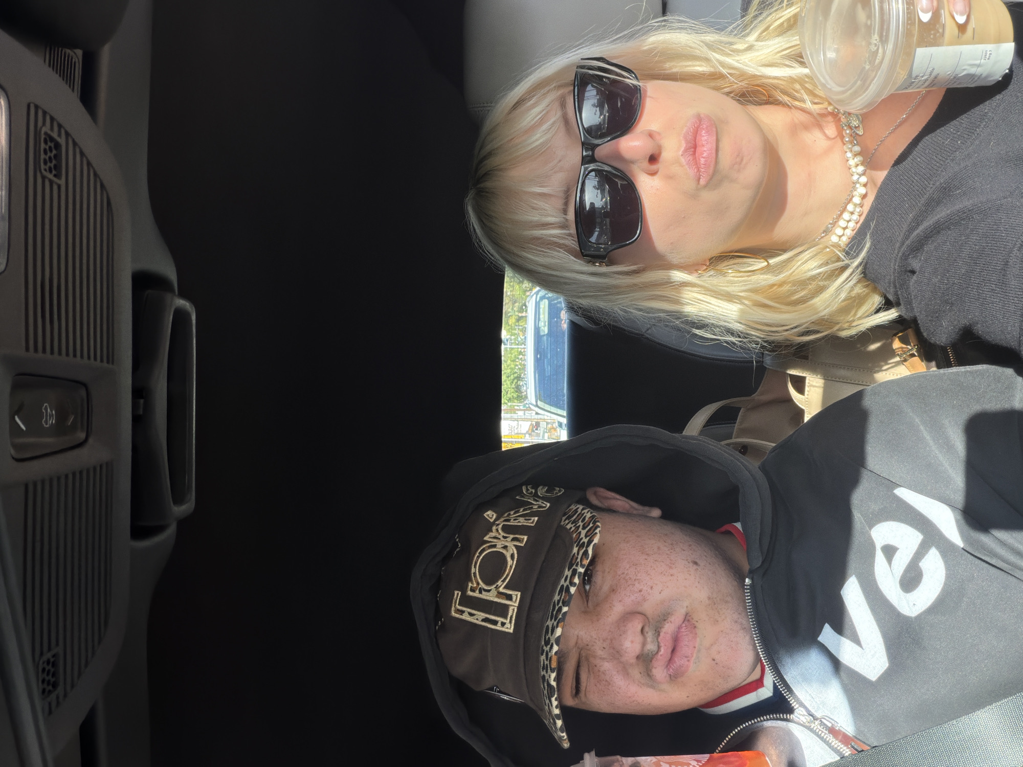 This media file shows two individuals inside a car, likely in West Hollywood, USA, during bright daylight. The image is oriented sideways, captured from a high angle looking down into the vehicle.
In the upper part of the frame is a blonde woman with shoulder-length hair, wearing black sunglasses and a black long-sleeved top. She has a multi-strand pearl necklace and a silver chain, and holds a clear plastic cup, resembling a Starbucks cold cup, containing a light brown beverage. Her expression is a slight pout. Sunlight casts a bright glow on her hair and face.
Below her is another person, wearing a dark hooded sweatshirt and a brown baseball cap with an animal print (leopard) brim. The cap features gold-colored, ornate text that appears to spell "Loud." This individual has visible freckles on their face and a relaxed, somewhat neutral expression with slightly parted lips. A red and white striped collar is visible beneath their hoodie. On the dark hoodie, white letters spelling "vel" are prominently visible, likely part of a larger word.
The interior of the car is dark, but details of the dashboard or door panel with vents and buttons are visible on the far left. The background visible through the car windows suggests a sunny, outdoor environment, possibly an urban or suburban street. The scene appears to be a casual moment during a drive or stop.