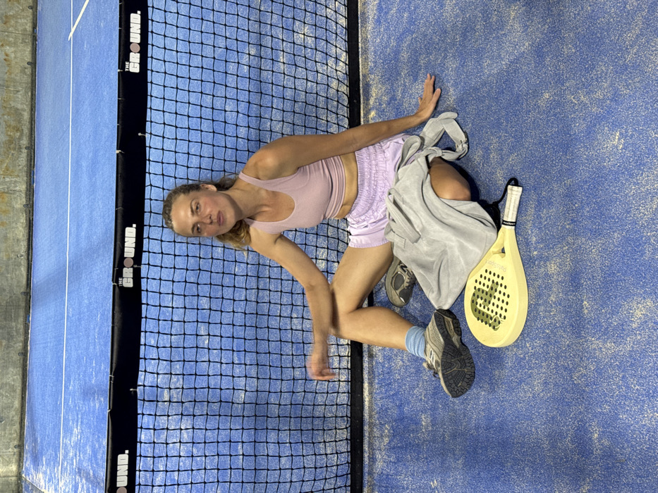 An adult female is depicted seated directly on a blue sports court in Pecatu, Indonesia. She wears a light pink athletic crop top and light purple athletic shorts, complemented by blue socks and grey athletic shoes. Her light brown hair is pulled back, and her facial expression is neutral with slightly pursed lips. Her right hand rests on the court surface, while her left leg is bent. Behind her is a black mesh net, typical of a padel court, featuring a black band at the top with "THE GROUND." printed in white text. A light grey fabric bag is positioned on the court next to her left leg. In front of her, a light yellow padel racket with a perforated face and a black and white grip rests on the court. The court surface is blue with visible white speckling, and a white boundary line is visible further back on the court. A light-colored wall with some texture is visible in the background above the court.