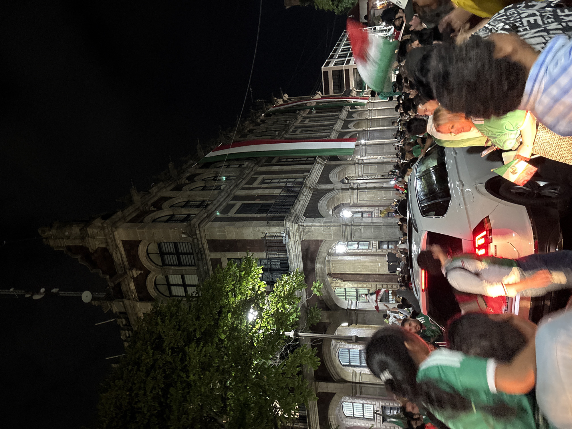 A nighttime street scene in Cuernavaca, Mexico, captures a large crowd gathered in front of a multi-story building adorned with large Mexican flags. The building, with its stone facade and arched windows, appears to be a historic structure. A white SUV is partially visible, surrounded by people who are holding more Mexican flags and appear to be celebrating or watching an event. The lighting is artificial, with streetlights illuminating the scene and casting shadows. The atmosphere suggests a festive or patriotic gathering.