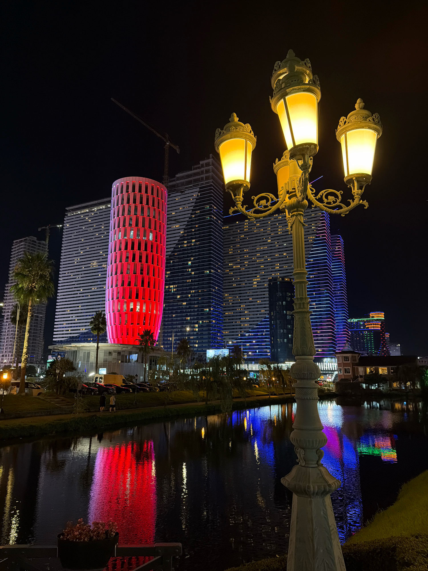 The image captures a nighttime scene in Batumi, Georgia, focusing on the waterfront area. Dominating the background are modern high-rise buildings, some illuminated with vibrant red and blue lights. One cylindrical building stands out with its red facade. In the foreground, a vintage-style lamppost with three illuminated globes is prominently featured. The lamppost is ornate and cast-iron, with decorative scrollwork. A calm body of water, likely a canal or a section of the Black Sea, reflects the colorful lights of the buildings and the lamppost. Palm trees are visible along the embankment, and the overall atmosphere suggests a lively urban environment at night. There are a few people walking on the embankment in the middle ground, and parked cars are visible along the street.
