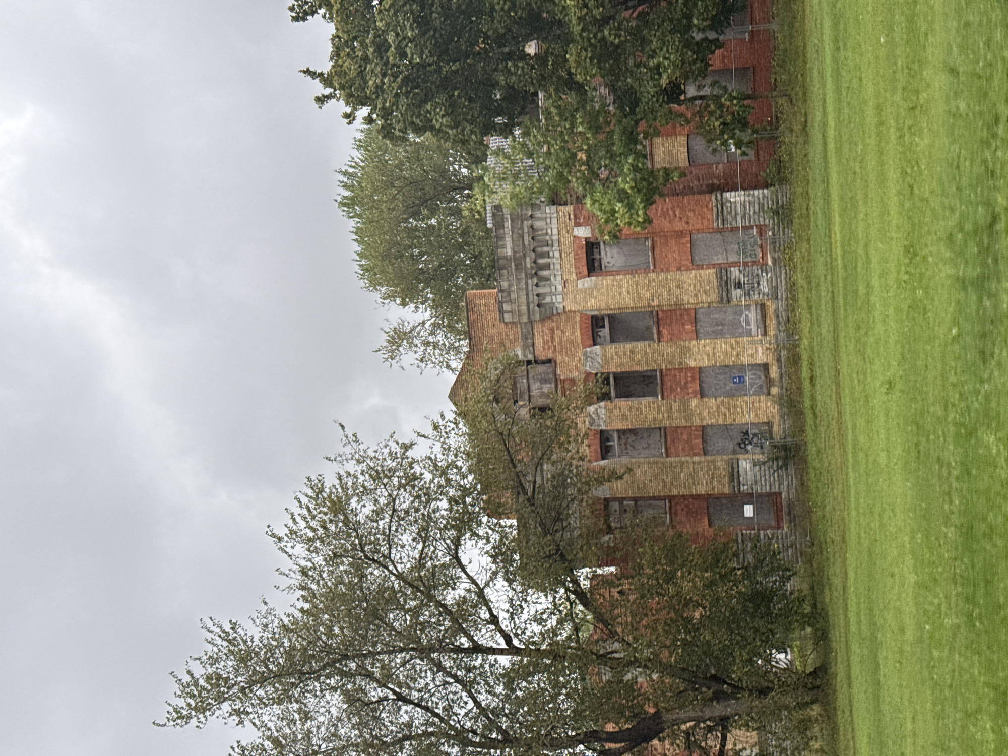 A low-angle, eye-level shot captures an imposing, abandoned brick building on a cloudy day. The building's facade features alternating bands of red and yellow brick. Many windows are boarded up with gray planks. Ornate stone detailing adorns the upper sections of the building. A mature tree with green foliage stands in front of the left side of the building, its branches partially obscuring the structure. Dense green trees fill the background. In the foreground, a manicured green lawn stretches across the scene. The image is presumed to be taken in Tallinn, Estonia.