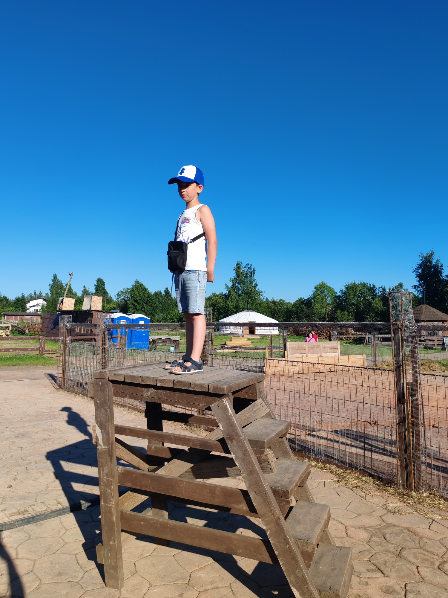 A young boy stands on a wooden platform at an outdoor petting zoo or farm in Lukashi, Russia. He is wearing a white tank top, denim shorts, a blue and white baseball cap, and sandals. He has a small black bag slung across his chest. The platform is elevated and has wooden steps leading up to it. The background shows a fenced-in area with various structures, including what appear to be animal enclosures, sheds, and possibly a yurt-like building. There are trees and a clear blue sky. The ground is paved with stone. The overall impression is of a sunny day at a rural recreational area.