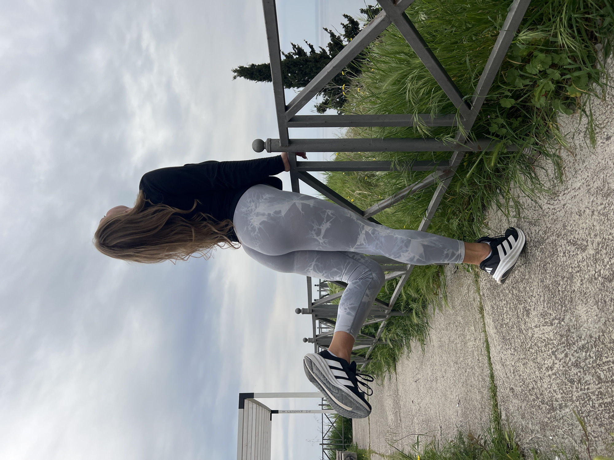 A woman is outdoors leaning against a metal railing with her back arched and head tilted back. She is wearing a black long-sleeved shirt and light grey patterned leggings with black and white Adidas sneakers. The background includes a cloudy sky, trees, grass, and part of a concrete walkway or path. The location is Prijevor, Montenegro.