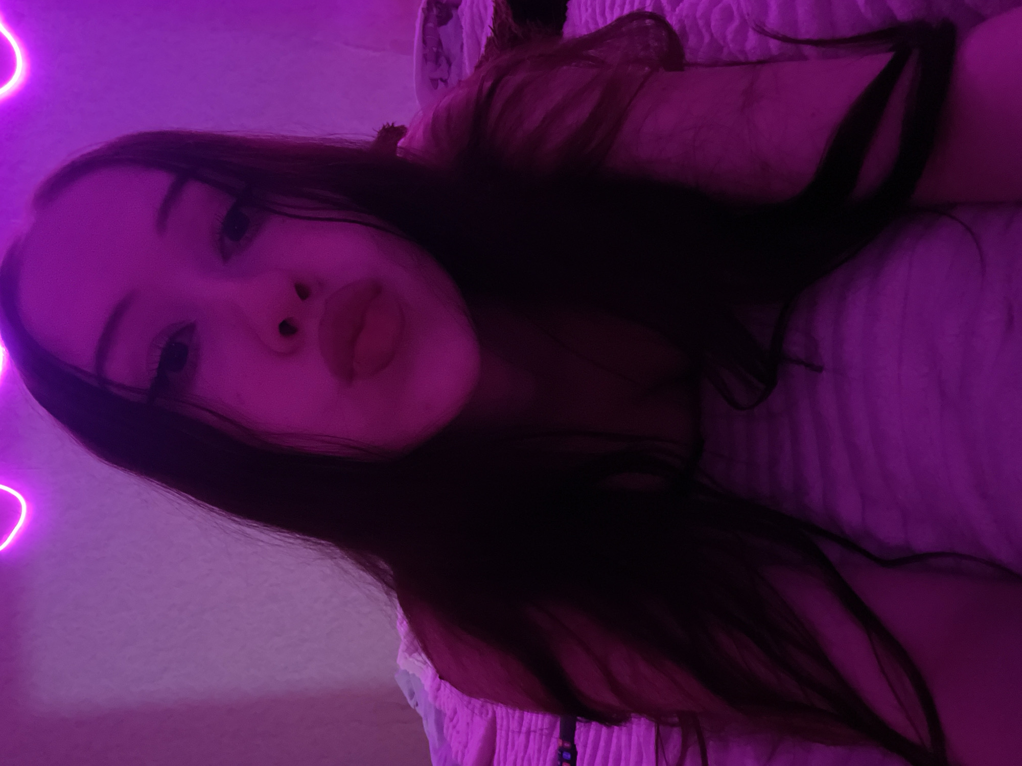 The image is a selfie of a young woman with long, dark hair, lying on her stomach in bed. She is looking at the camera with her lips puckered. The room is lit with pink neon lights, which cast a purple glow over the scene. The bed appears to be covered with a textured blanket. The background shows a pink wall with more neon lights. There are no identifiable landmarks or contextual clues that would allow for the determination of the location within Votkinsk, Russia.