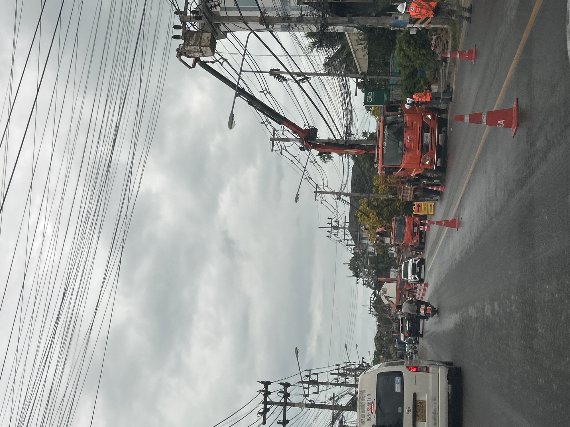In the city of Ko Samui District, Thailand, a street scene unfolds under an overcast sky, with numerous overhead power lines crisscrossing the frame. The road surface appears wet, suggesting recent rain. A white van is parked on the left side of the road. Further down the street, a motorcyclist rides away from the camera.
Several construction vehicles and workers are present. An orange crane truck with a bucket lift is positioned on the right side of the road, with a worker in the bucket attending to the power lines. Another orange truck is parked in front of it. Two workers in high-visibility vests are standing near the orange truck, and another worker in a similar vest is visible further down the road on the right. Traffic cones are placed along the road, indicating ongoing work or a closed lane. Trees and buildings line the right side of the street.