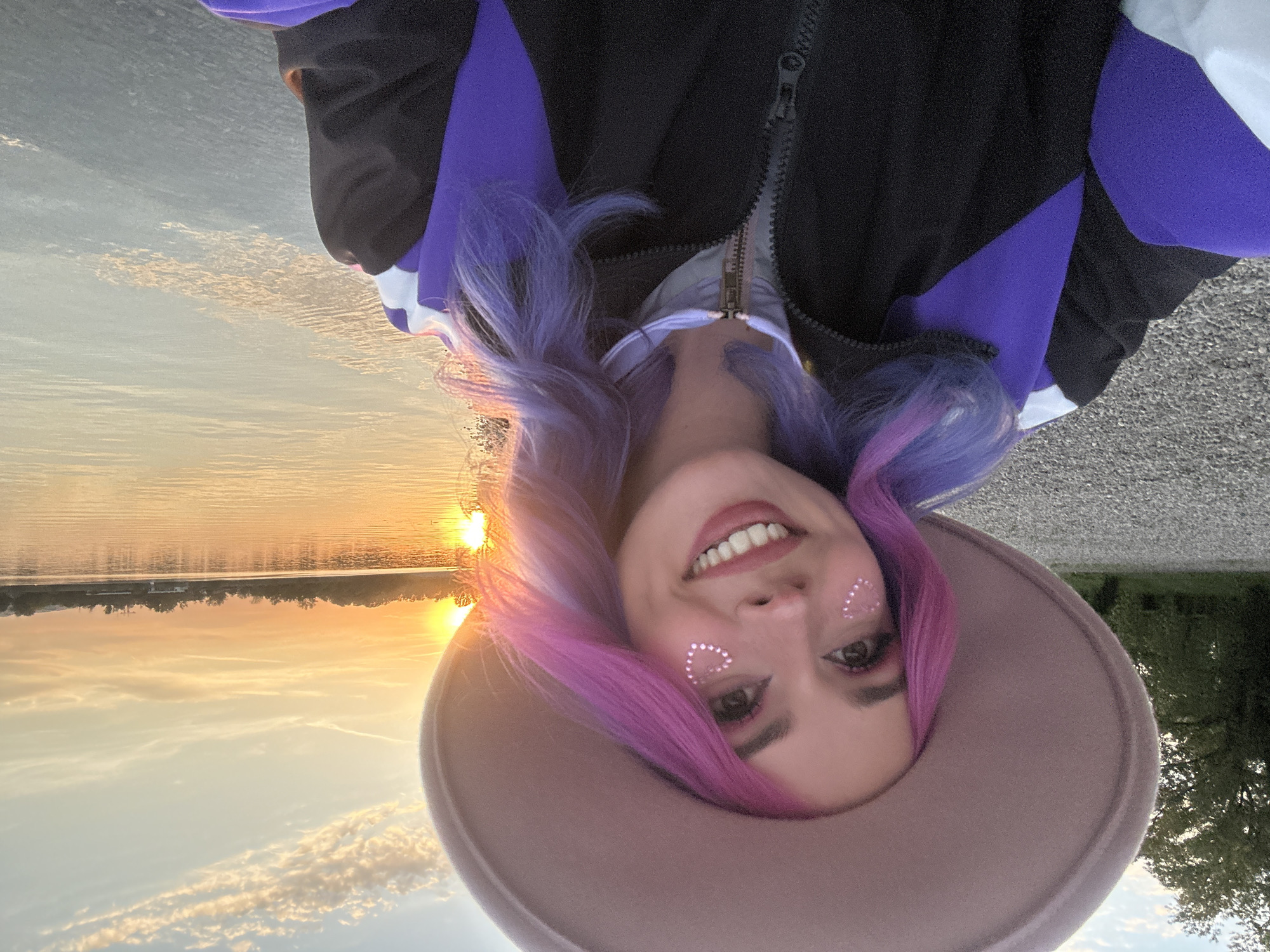 A close-up outdoor portrait captures a smiling young woman during sunset in the City of Zagreb, Croatia. She is looking directly at the camera with a joyful, broad smile, showing her teeth. Her long, wavy hair is dyed in striking shades of purple and pink. She wears a light brown or beige wide-brimmed hat that frames her face. Distinctive glittery, heart-shaped decals adorn her cheeks. Her attire includes a black, purple, and white athletic or casual jacket with a visible zipper.
The background features a serene sunset over a calm body of water, likely a lake or wide river, such as Jarun Lake or the Sava River. The sun is low on the horizon, casting a warm golden and orange glow across the water and sky. A distant treeline is visible on the far bank of the water. The sky above is clear with some scattered, soft clouds. The immediate foreground, beneath the woman, appears to be a paved or gravel surface. The image conveys a cheerful and peaceful mood, characteristic of the golden hour. No visible text is present in the media.