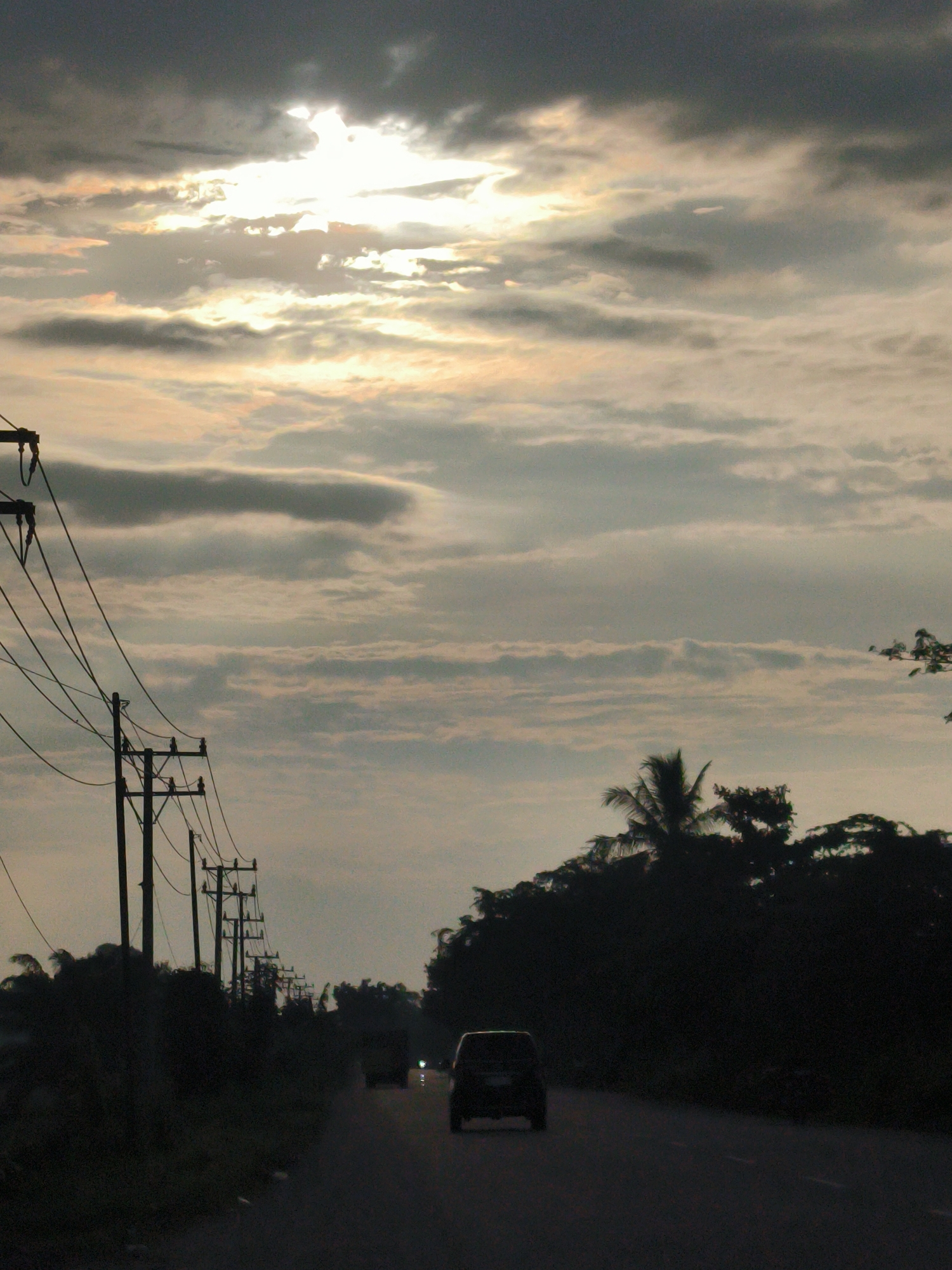 The image depicts a road in Cotentung, Indonesia, at dusk. The sky is filled with dramatic clouds, with the sun breaking through in the upper left quadrant, casting a bright, hazy light. On the left side of the road, a line of utility poles with overhead wires stretches into the distance. The right side of the road is bordered by a dense line of dark trees and palm fronds. Two vehicles are visible on the road: a dark SUV in the foreground, and another vehicle further back, with its headlights illuminated. The overall impression is of a journey on a darkening rural road with a striking sky.