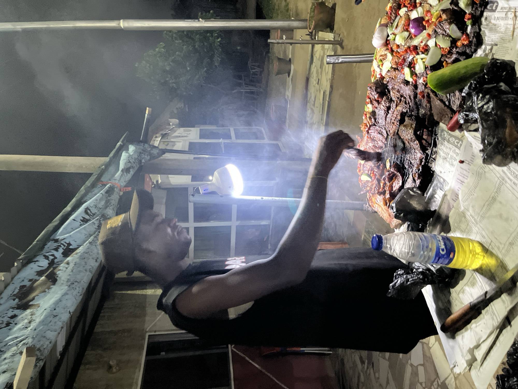 A nighttime outdoor street food stall in Abuja, Nigeria, features a man in a black sleeveless shirt and a brown baseball cap preparing grilled meat. He is standing behind a large display of barbecued meat, garnished with red chilies, lime wedges, cucumber slices, and red onions. A bottle of Citron-branded yellow liquid and a knife with a wooden handle are on the table in front of the meat. The stall is covered by a tarp supported by wooden beams. A bright, portable light illuminates the scene, casting shadows and creating visible smoke from the cooking meat. The background shows a building with windows and some outdoor seating. The ground is paved with mosaic tiles.