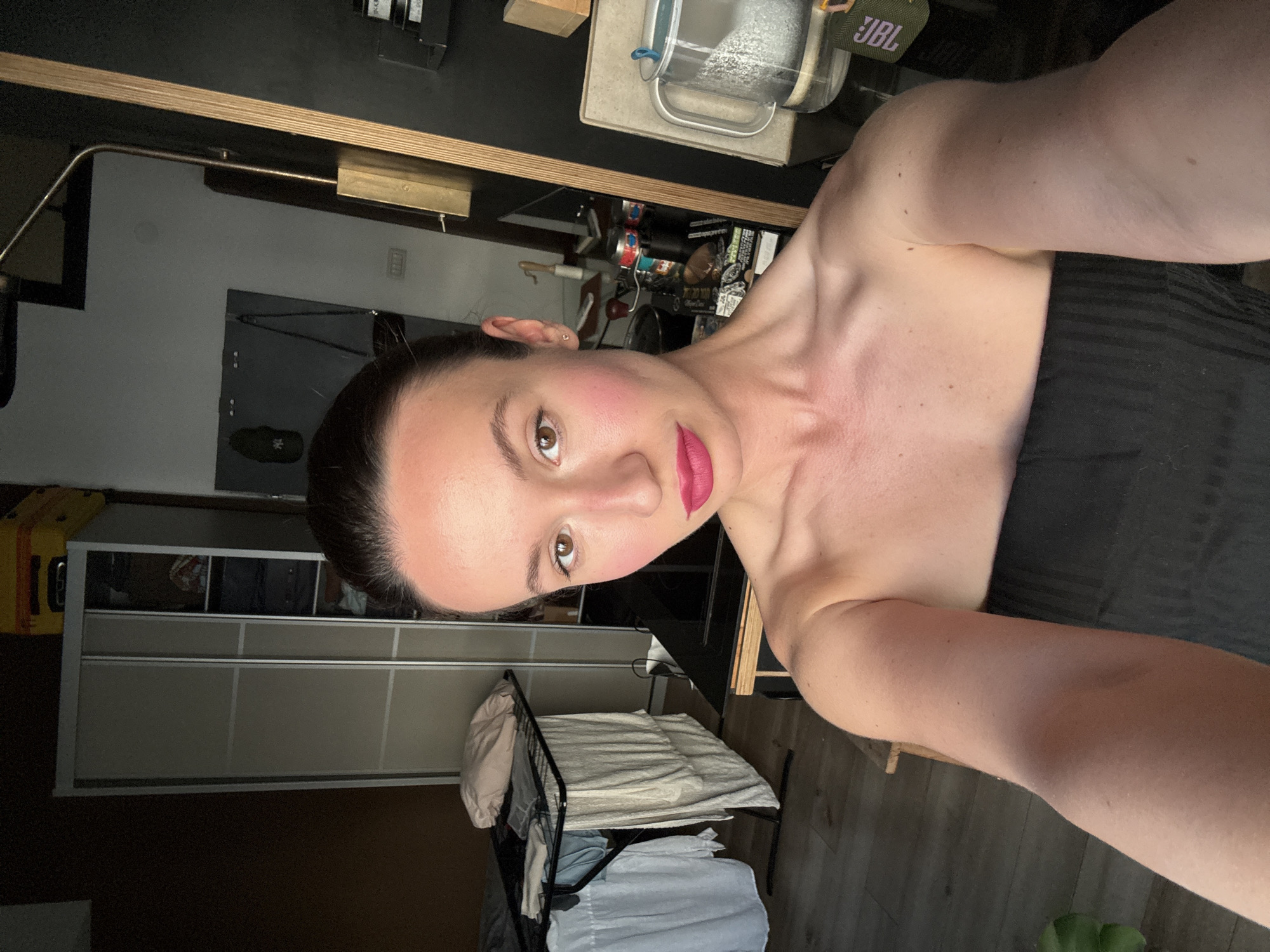 A selfie of a woman with fair skin, dark hair pulled back, wearing pink lipstick and blush, and a strapless dark-colored top. She is holding the camera at arm's length, tilted slightly.
Behind her is a room with a wardrobe/closet on the left and a drying rack with white and blue clothes on the bottom. A desk with various items, including a JBL speaker and what appears to be cleaning supplies, is visible in the background. A light fixture is mounted under a shelf above the desk. The setting is indoors. I cannot determine the specific location in Tel-Aviv, Israel, based on this image alone.