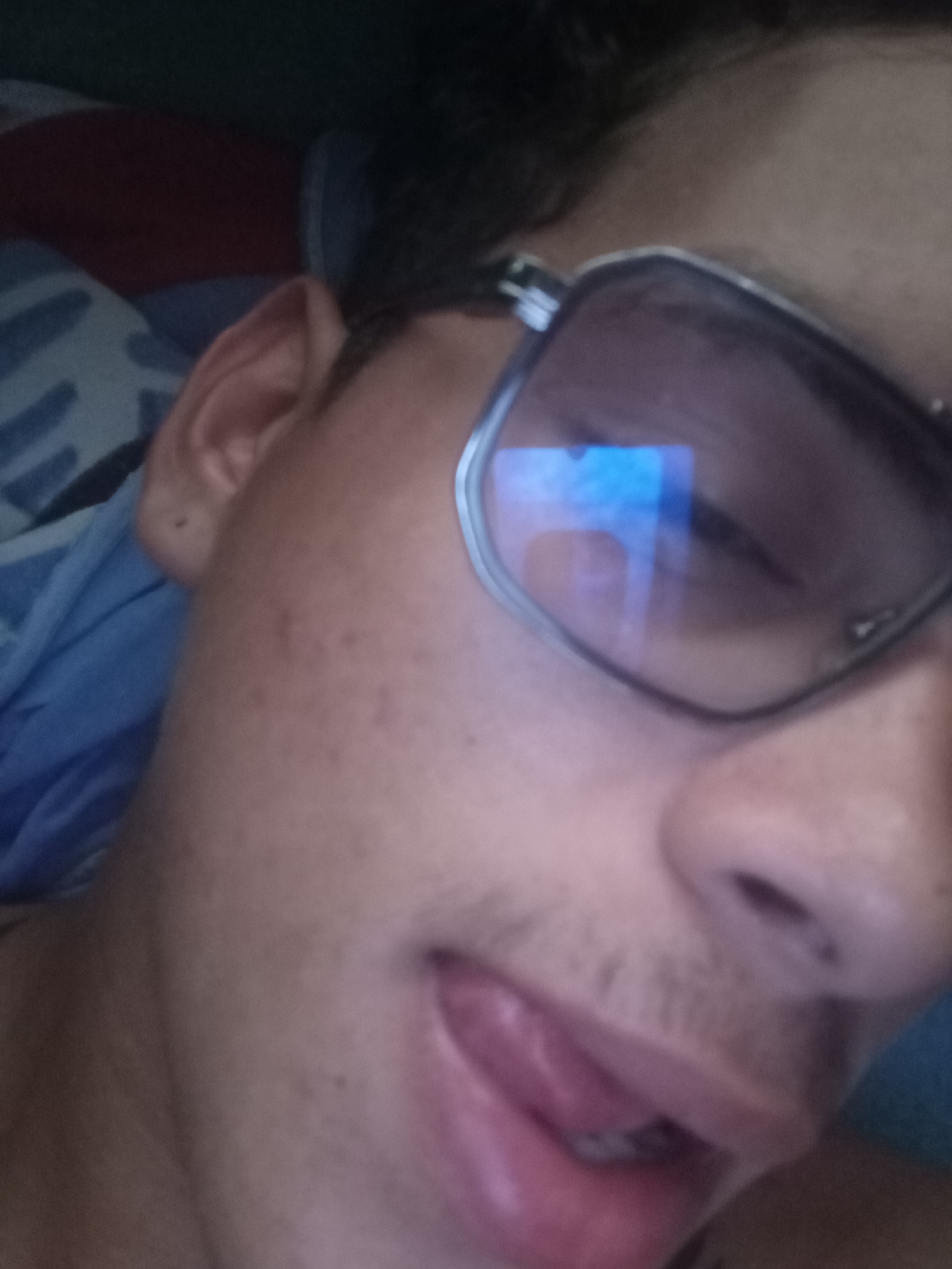 The image shows a person wearing glasses. The person's tongue is slightly sticking out. The background includes blue and red objects. The location is unknown and cannot be determined from the image.